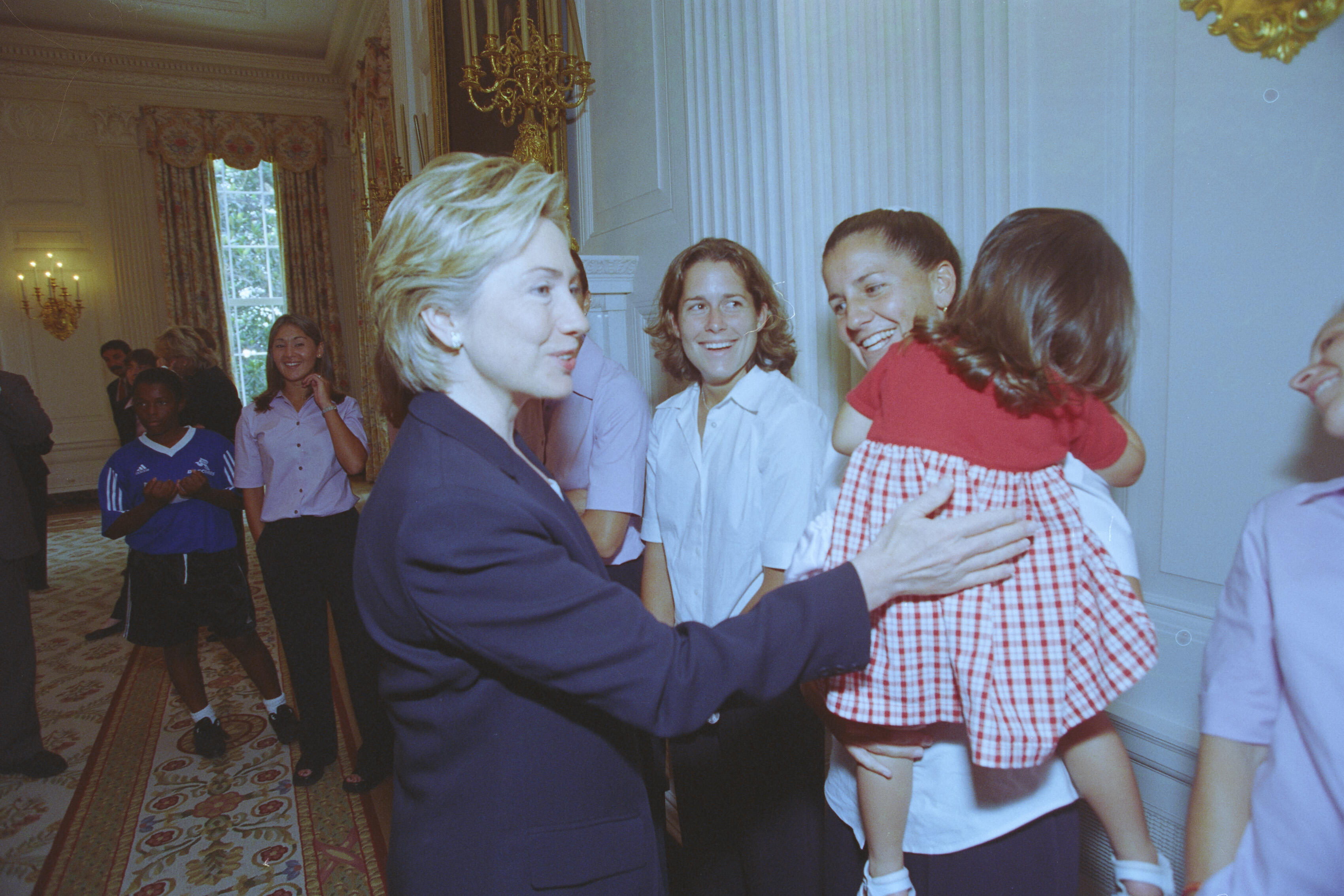 A medium shot captures First Lady Hillary Clinton smiling as she interacts with a group of women in a formal White House room. She places her hand on the back of a young girl wearing a red and white gingham dress, who is being held by a smiling woman in a white short-sleeved shirt. Other smiling women in matching light pink shirts look on, while a young girl in a blue sports jersey stands further back in the hallway.