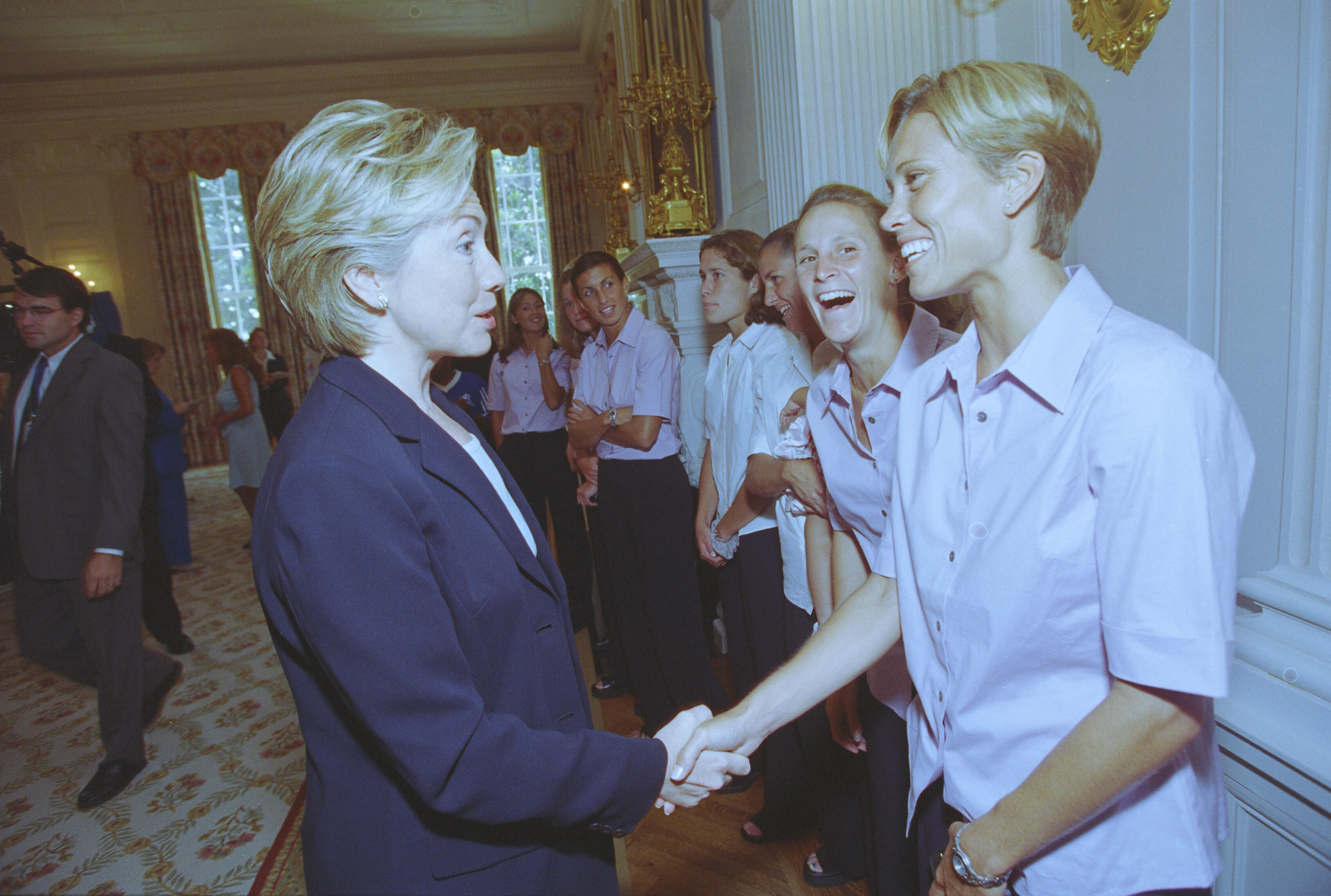 A medium shot captures First Lady Hillary Clinton from a side profile as she shakes hands with a smiling woman with short blonde hair. Behind her, another woman with her blonde hair pulled back laughs joyfully, both wearing matching light pink short-sleeved button-down shirts. A line of other smiling women stands against the white paneled wall of an elegant room, while a man in a grey suit walks in the background on the far left.