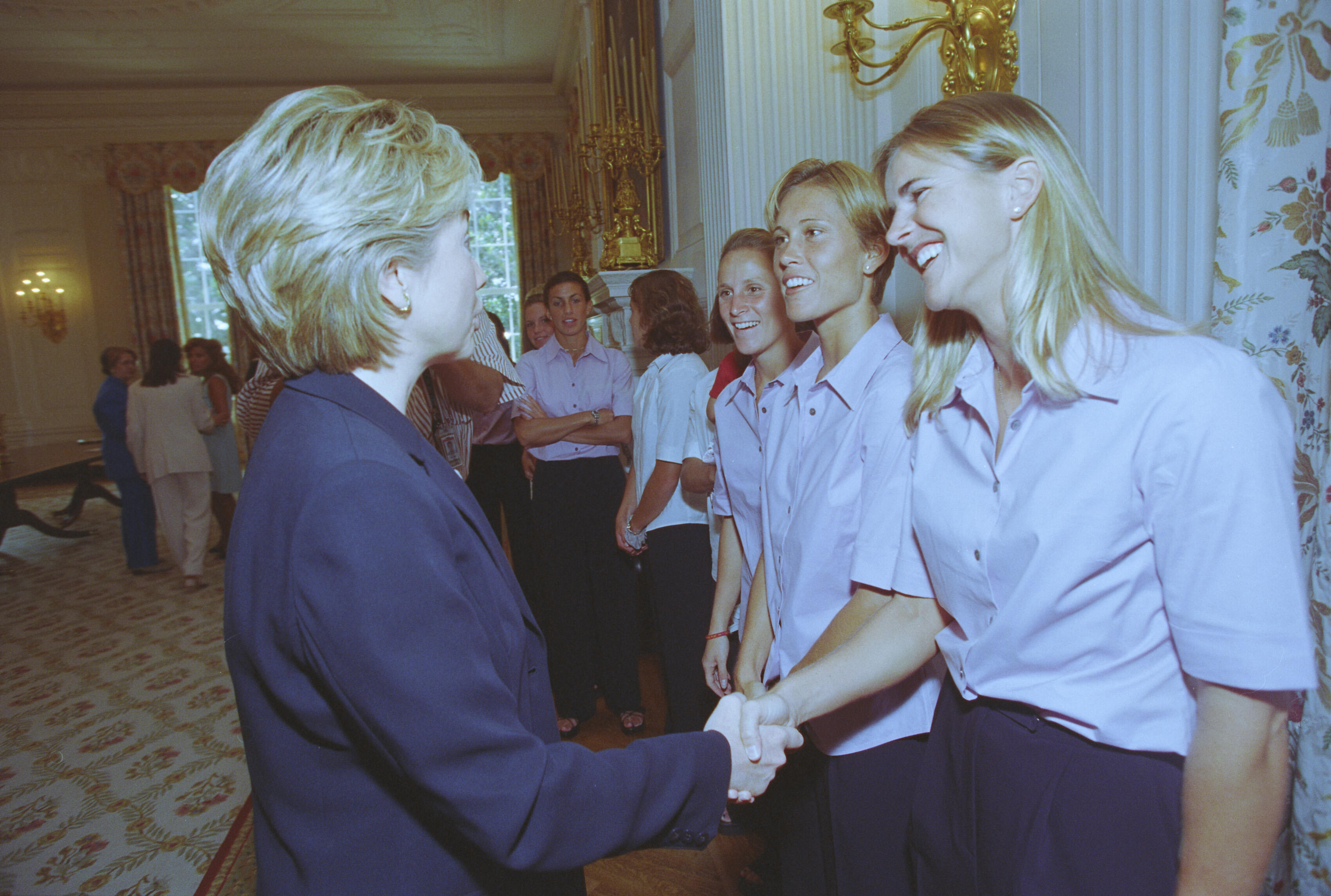 A medium shot captures First Lady Hillary Clinton from a side profile as she shakes hands with a laughing blonde woman standing at the front of a line. She and the other women behind her wear matching light pink short-sleeved button-down shirts and dark pants. The greeting takes place in a brightly lit room next to a white wall with an ornate gold sconce and floral-patterned curtains on the far right.