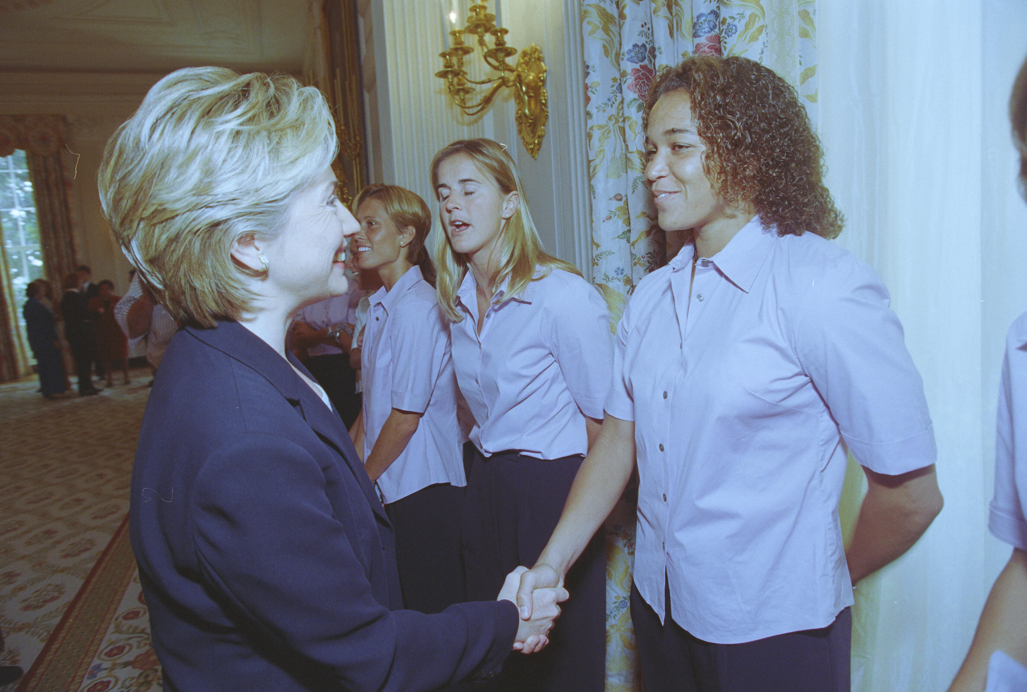 A medium shot captures First Lady Hillary Clinton from a side profile as she smiles and shakes hands with a smiling woman with curly hair. Two other women stand behind her in line, all wearing matching light pink short-sleeved button-down shirts and dark pants. The interaction occurs near a white wall decorated with an ornate gold sconce and floral-patterned curtains on the right.