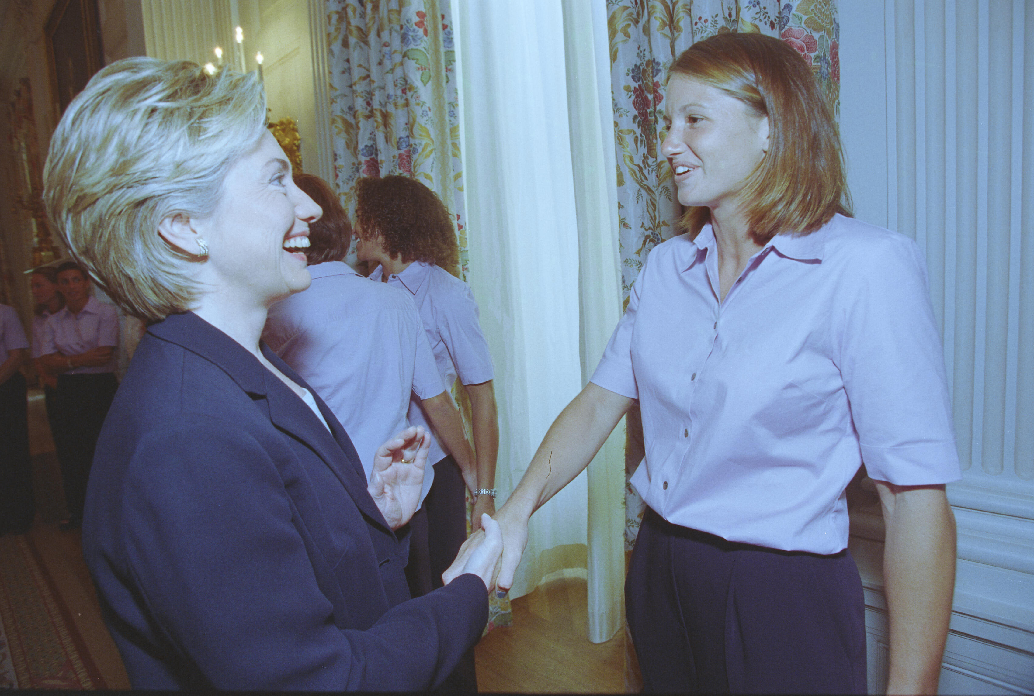 A medium shot captures First Lady Hillary Clinton from a side profile as she laughs and shakes hands with a smiling woman with shoulder-length brown hair. The woman wears a light pink short-sleeved button-down shirt and dark pants, matching the other women visible in the background. They stand in an elegant room next to white walls and tall windows draped with floral-patterned curtains.