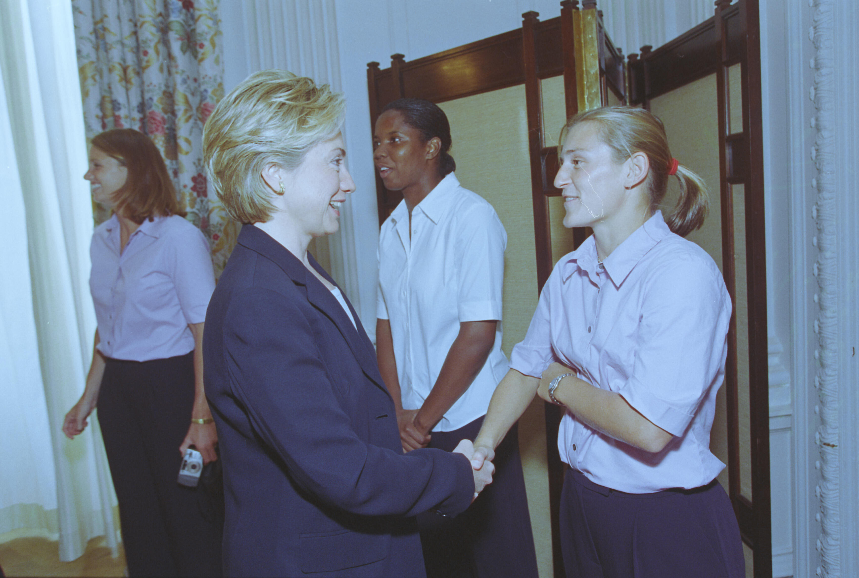 A medium shot captures First Lady Hillary Clinton from a side profile as she smiles and shakes hands with a blonde woman with her hair in a ponytail. Behind them stands a Black woman in a white button-down shirt, and another woman in a light pink shirt is visible on the far left. The interaction takes place in front of a folding privacy screen and a white wall, with floral-patterned curtains on the left side of the room.