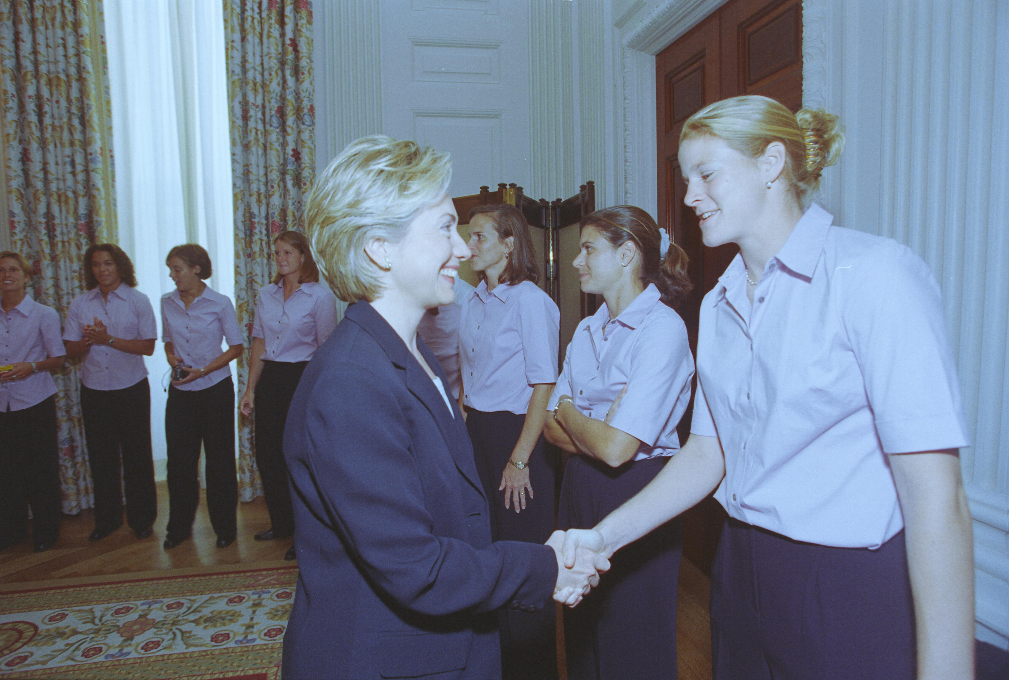 A medium shot captures First Lady Hillary Clinton from a side profile as she smiles and shakes hands with a smiling blonde woman with her hair in a bun. On the left, several other women stand in a line, watching the interaction, while two women stand close behind the blonde woman. The group wears matching light pink short-sleeved button-down shirts and dark pants, gathered in a brightly lit room featuring a patterned rug and floral-patterned curtains on the left.