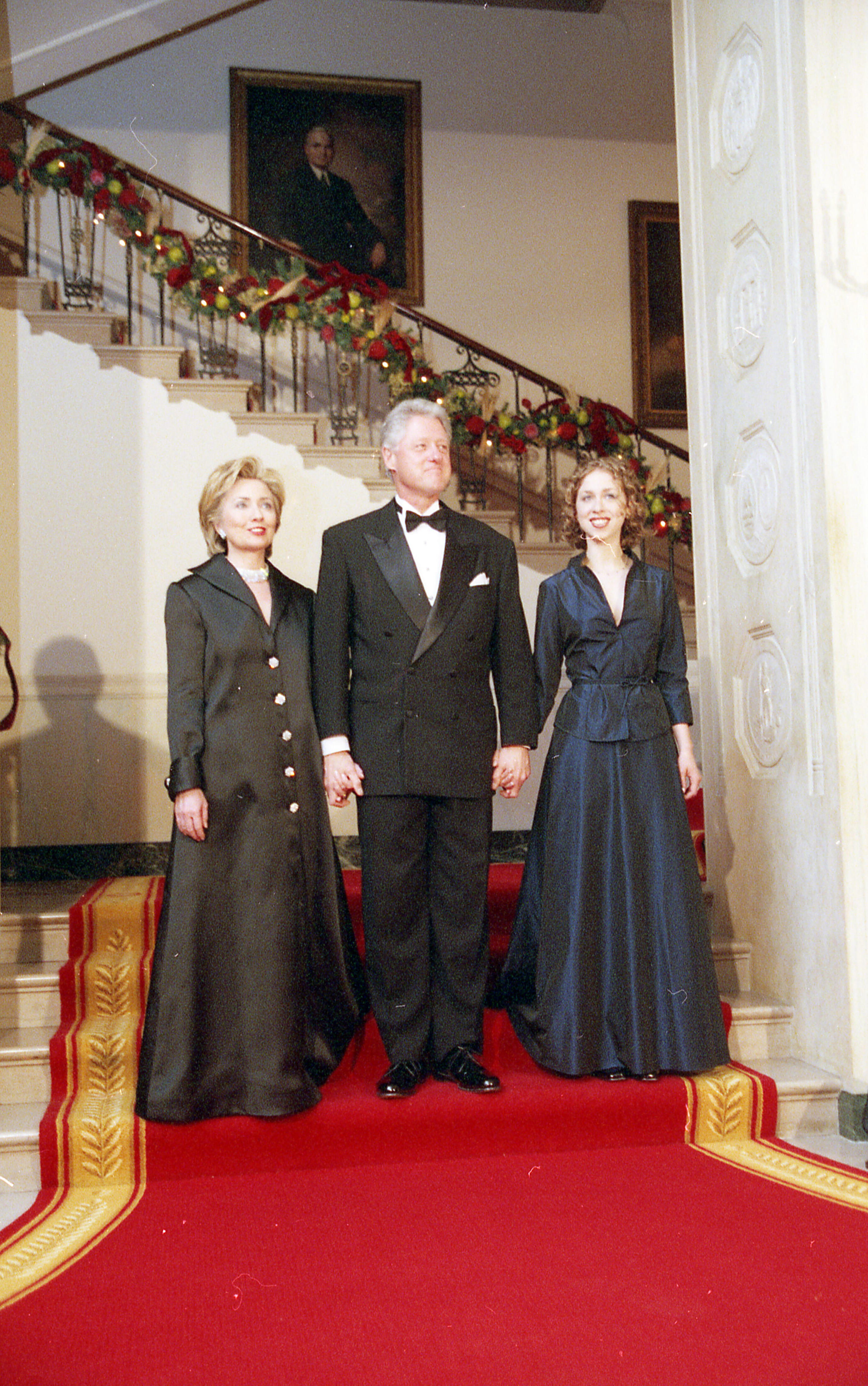 President Clinton, Hillary Rodham Clinton, and Chelsea Clinton stand together in the doorway of the East Room. The Grand Staircase decorated with Christmas Garland can be seen in the background.