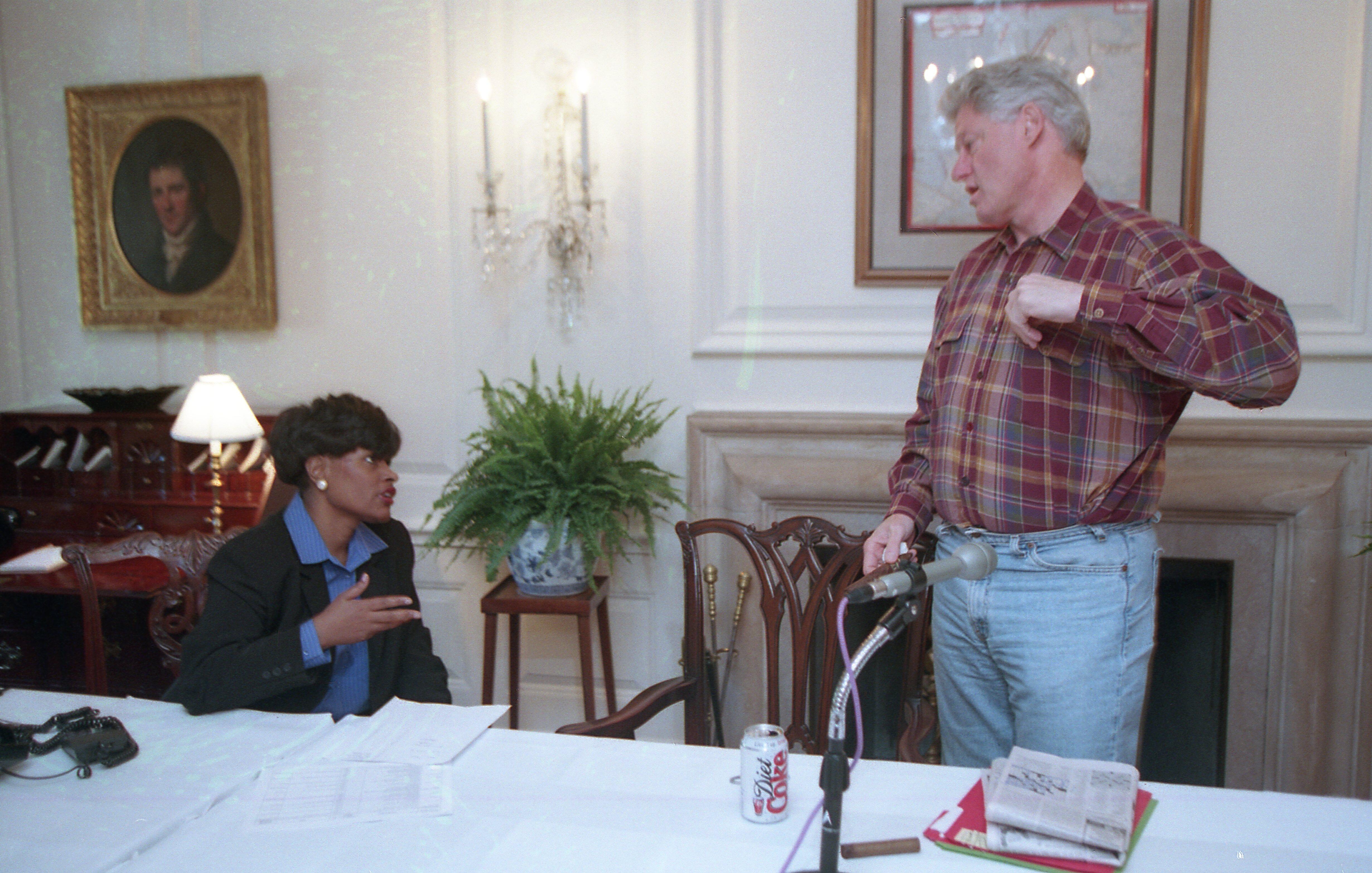 President Clinton Stands in blue jeans. Minyon Moore sits behind a table covered in white table cloth.