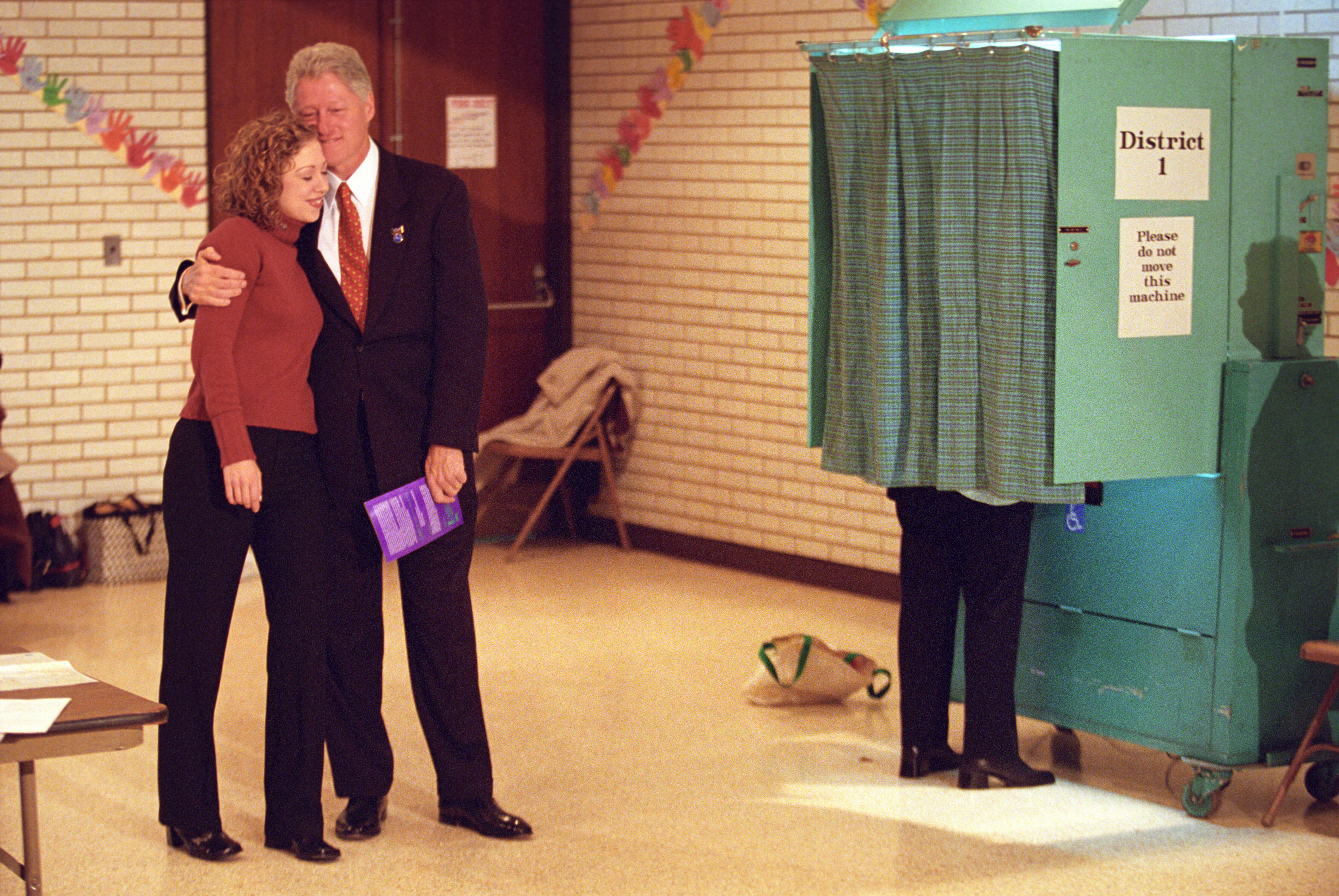 President Clinton hugs Chelsea Clinton outside of a aqua color voting booth. The booth's curtain is closed. Both are standing. President Clinton wears a suit.