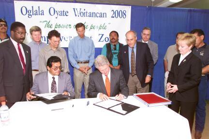 President Clinton sits a round table. He signs a document. Community and business leaders stand around President Clinton.