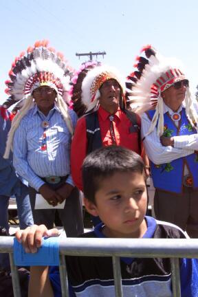 Three Native Americans in ceremonial feathered headdresses and a young boy stand and listen a signing ceremony at the Pine Ridge Reservation in South Dakota. The boy stands behind a crowd control rail.