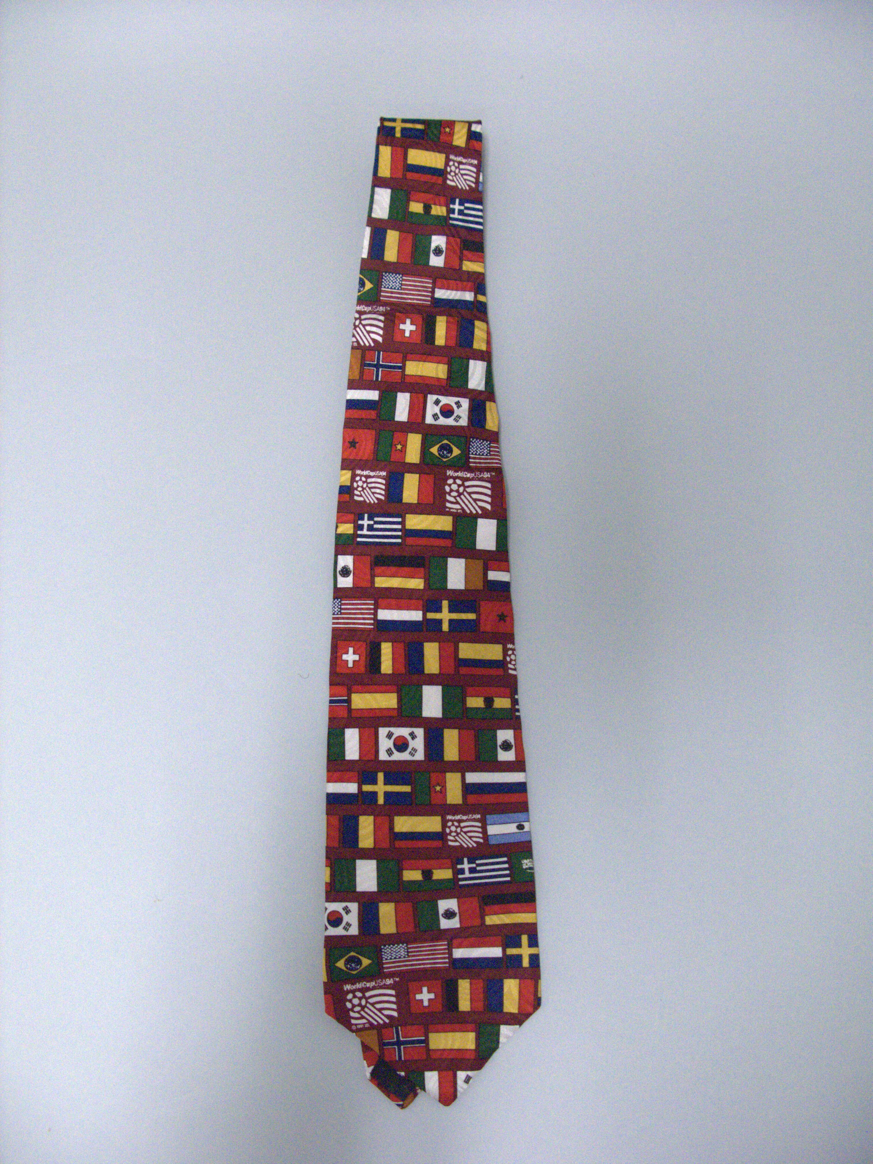 A necktie with a repeating grid pattern of international flags is laid vertically against a plain light grey background. The colorful pattern includes the national flags of various countries like the United States, Greece, Switzerland, and South Korea, interspersed with the official "WorldCupUSA94" soccer logo. The tie narrows toward the top and terminates in a pointed tip at the bottom.