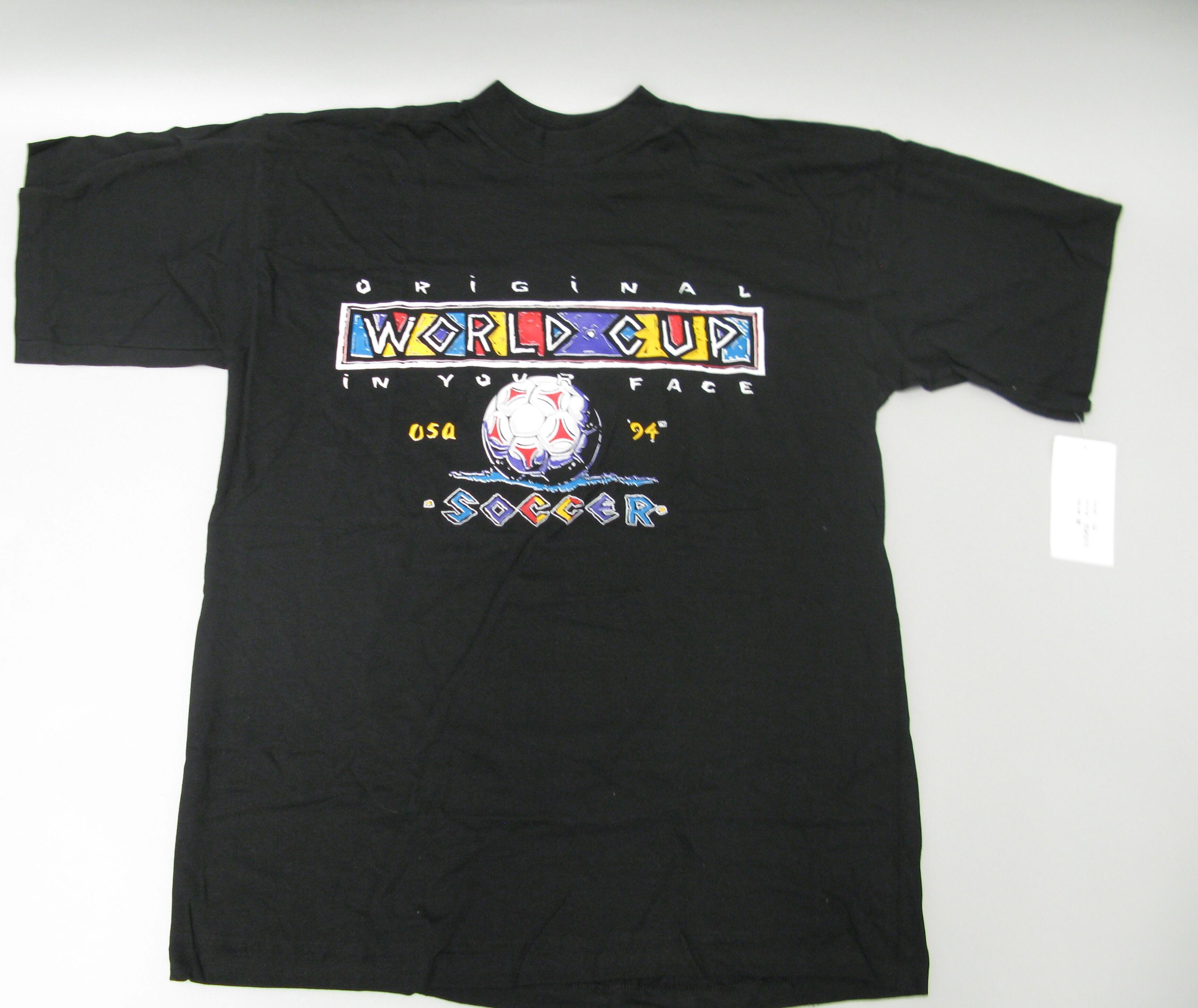 A black crewneck t-shirt is laid flat against a light grey surface, featuring a colorful, stylized 1990s graphic on the front. At the top of the graphic, the word "ORIGINAL" is printed in spaced white letters above a rectangular box containing the words "WORLD CUP" in an artistic font with shades of purple, yellow, and blue. Below this box, the phrase "IN YOUR FACE" is printed in white letters, followed by a central illustration of a soccer ball splashes into blue water. The yellow text "USA 94" flanks ....