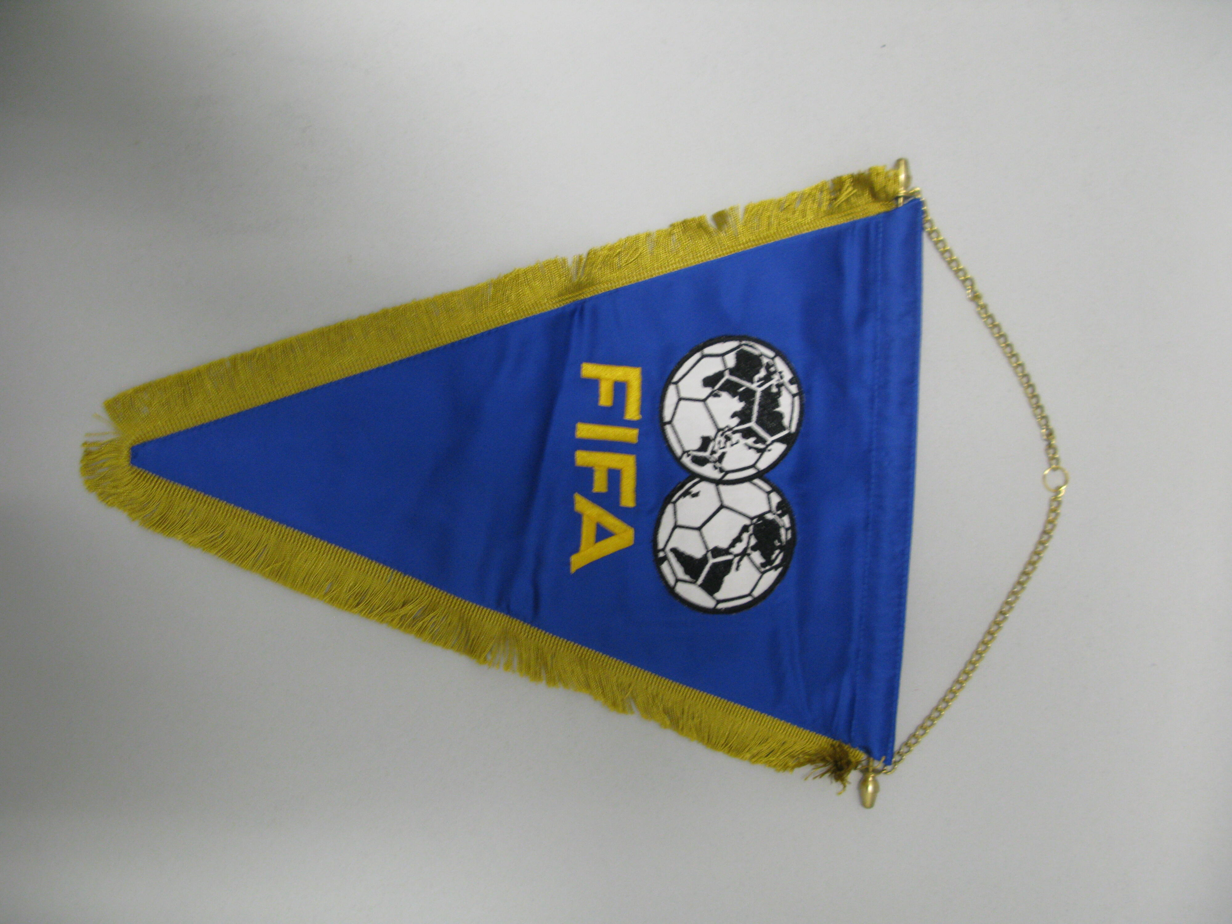 A triangular blue fabric pennant is displayed vertically against a plain light grey background, suspended by a thin gold chain attached to a horizontal metal rod at the top. The pennant features the word "FIFA" printed in solid yellow block letters across the center. Directly above the text, two overlapping soccer balls are illustrated, with the continents of a world map filled in black on their panels. The two sides of the triangle are trimmed with thick, golden-yellow fringe that tapels down to a point...