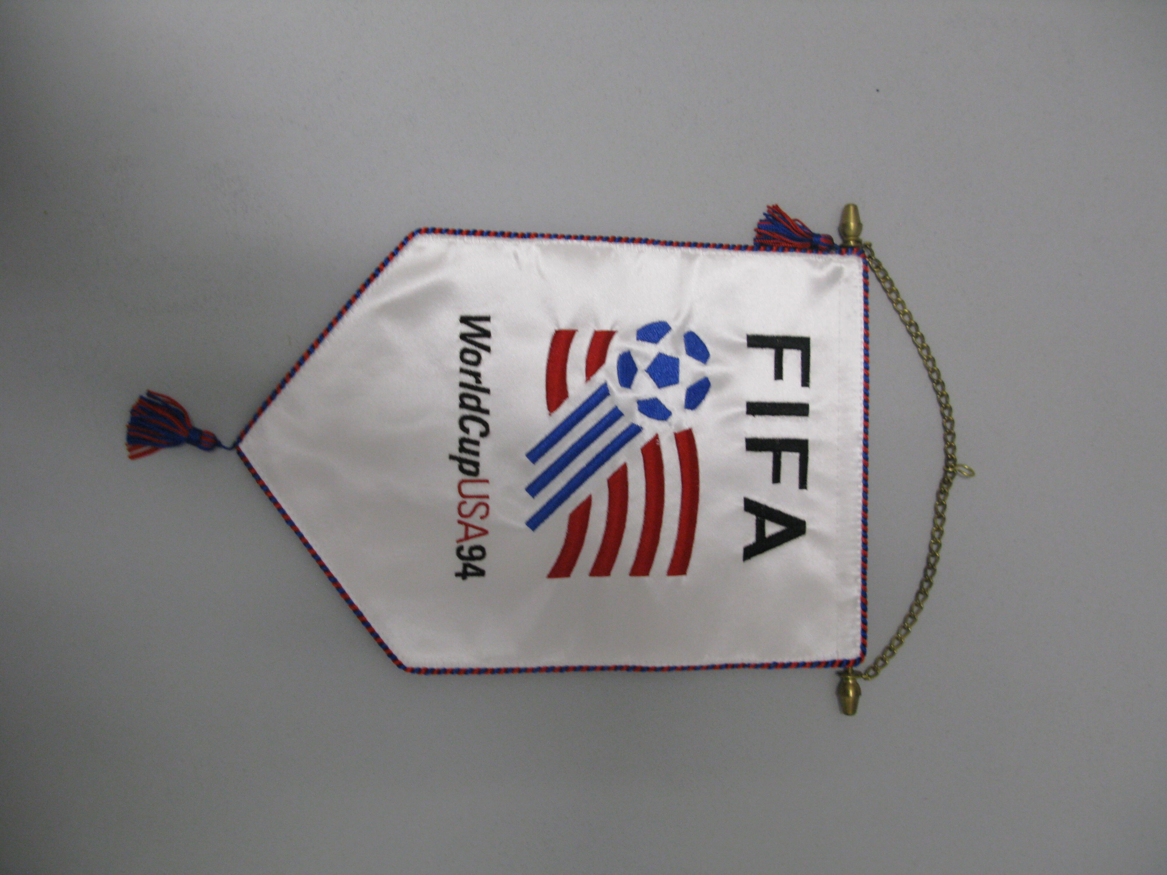 A white satin-like souvenir pennant hangs vertically against a plain light grey background, suspended by a thin gold chain attached to a horizontal metal rod at the top. The pennant features the word "FIFA" printed in large, black block letters at the top. Below this, the official 1994 FIFA World Cup logo is displayed, featuring a blue soccer ball with trailing red and blue stripes resembling a flag. The text "WorldCupUSA94" is printed underneath the logo in black and red. 