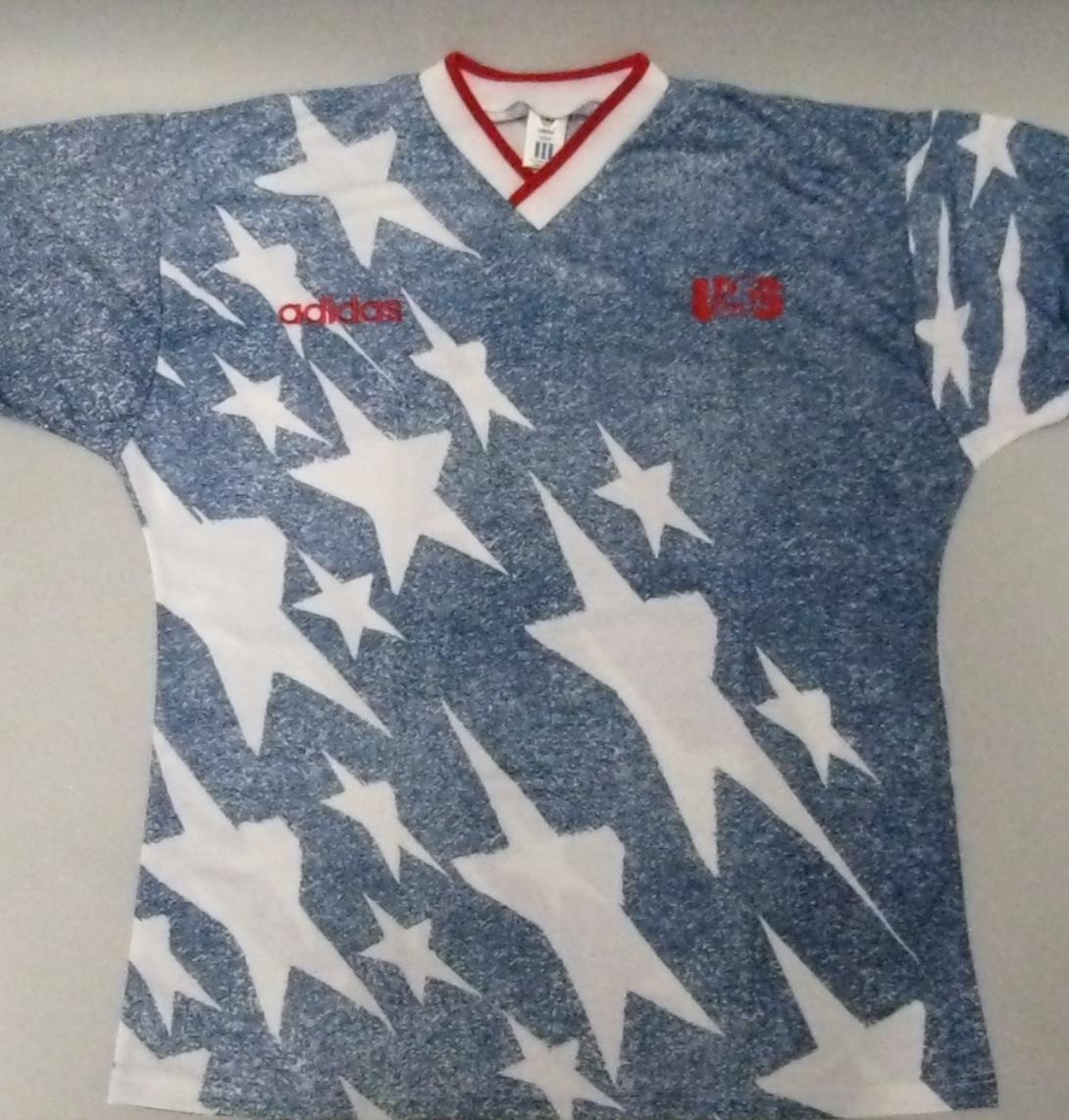 A short-sleeved soccer jersey with a blue and white faux-denim texture is laid flat against a plain light grey background. The jersey features a bold, scattered pattern of large and small white stars cascading diagonally down the left side and across the right sleeve. It has a white V-neck collar and white sleeve cuffs, both trimmed with a thin red stripe. On the right chest, the Adidas wordmark is embroidered in red, while the letters "US" are embroidered in red on the left chest.