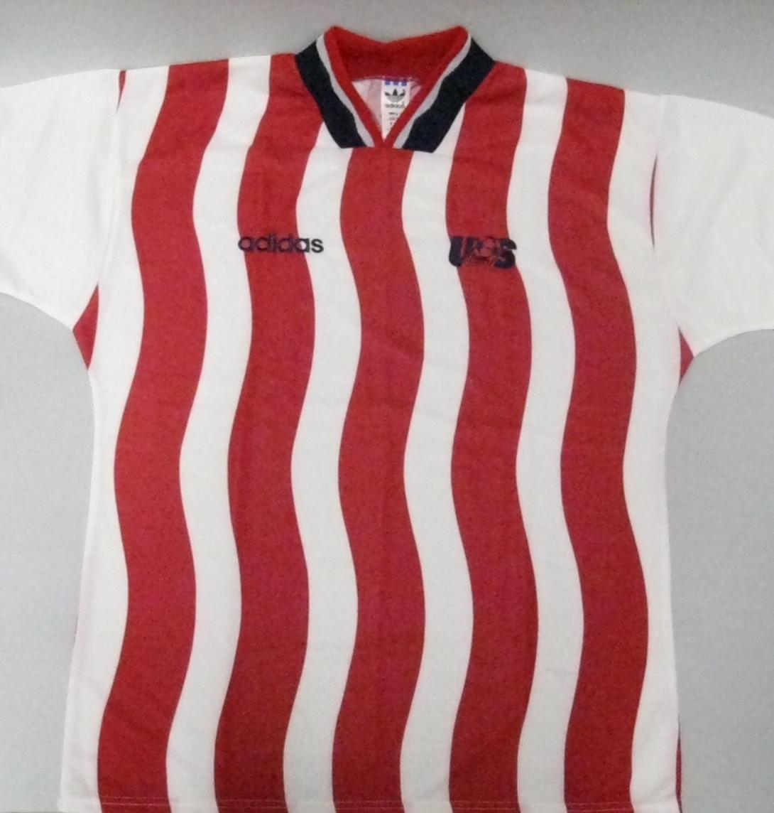 A short-sleeved soccer jersey featuring vertical red and white wavy stripes on the torso is laid flat against a light grey background. The sleeves are solid white and finished with dark blue and red striped cuffs. The jersey has a dark blue collar with a red and white inner V-neck trim. On the right chest, the Adidas logo is embroidered in dark blue, while a stylized "US" logo is embroidered on the left chest.