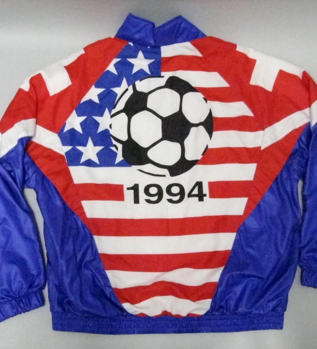 The back view of a vintage tracksuit jacket is laid flat, featuring a bold red, white, and blue graphic design celebrating the 1994 World Cup. The center panel displays a large black and white soccer ball set against a background mimicking the American flag, with white stars on a blue field to the left and horizontal red and white stripes across the rest of the panel. The year "1994" is printed in large black numbers directly beneath the soccer ball. The jacket has solid blue sleeves.