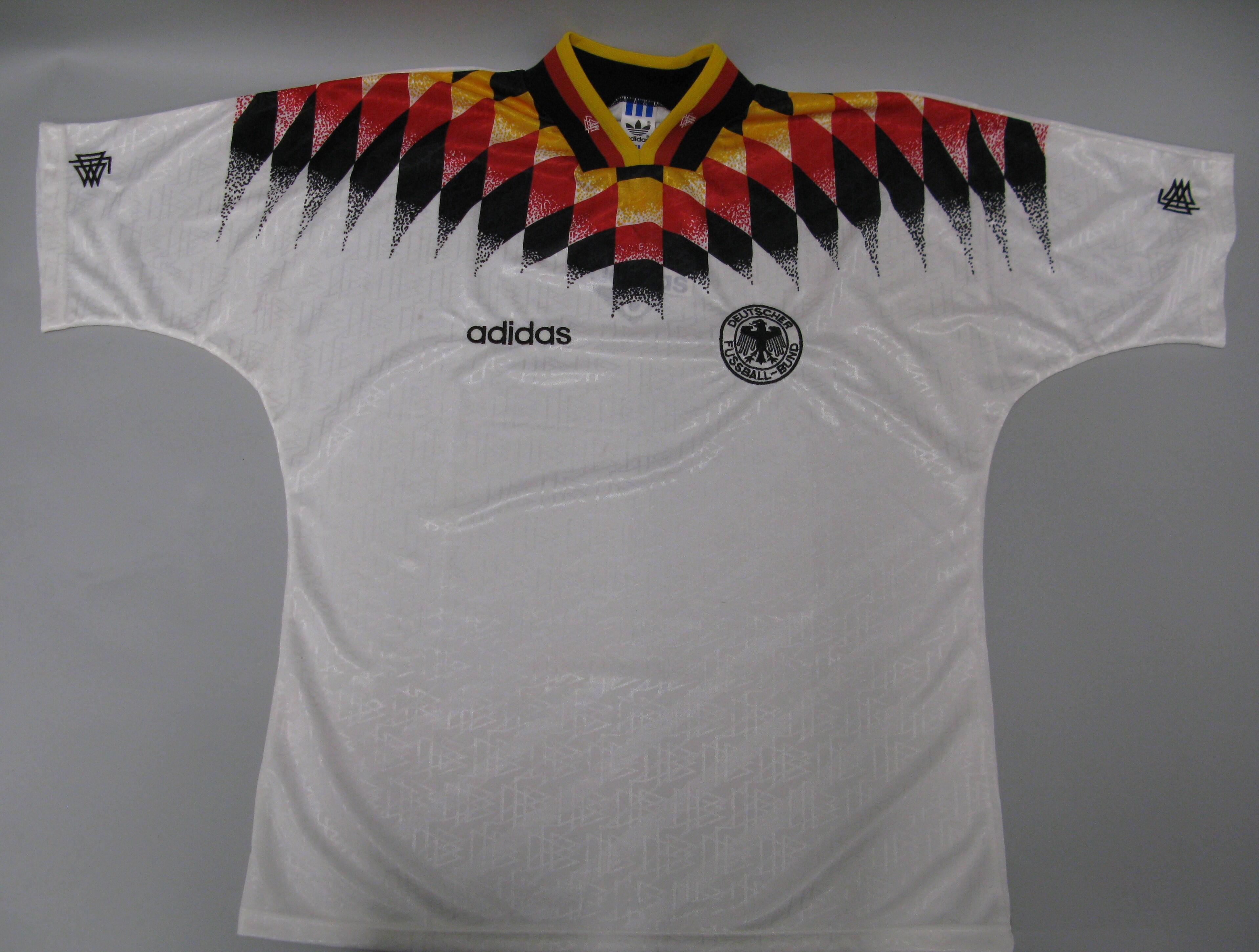 A white short-sleeved football jersey featuring a subtle, repeating geometric jacquard pattern woven into the fabric is laid flat against a light grey background. A bold graphic chevron pattern in the colors of the German flag—yellow, red, and black—stretches across the chest, shoulders, and collar area in a pixelated diamond gradient design. The jersey has a black collar trimmed with yellow and red stripes. The black Adidas wordmark is printed on the right chest, and the circular "Deutscher Fussball-Bund" 