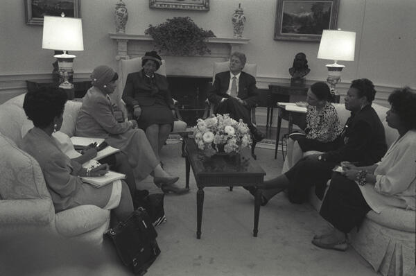 Black and white photograph. President Clinton sits in the Oval Office with Black Women's Leadership including: Maya Angelou, Dorothy Height, Delores Tucker, Alexis Herman, and Maggie Williams,