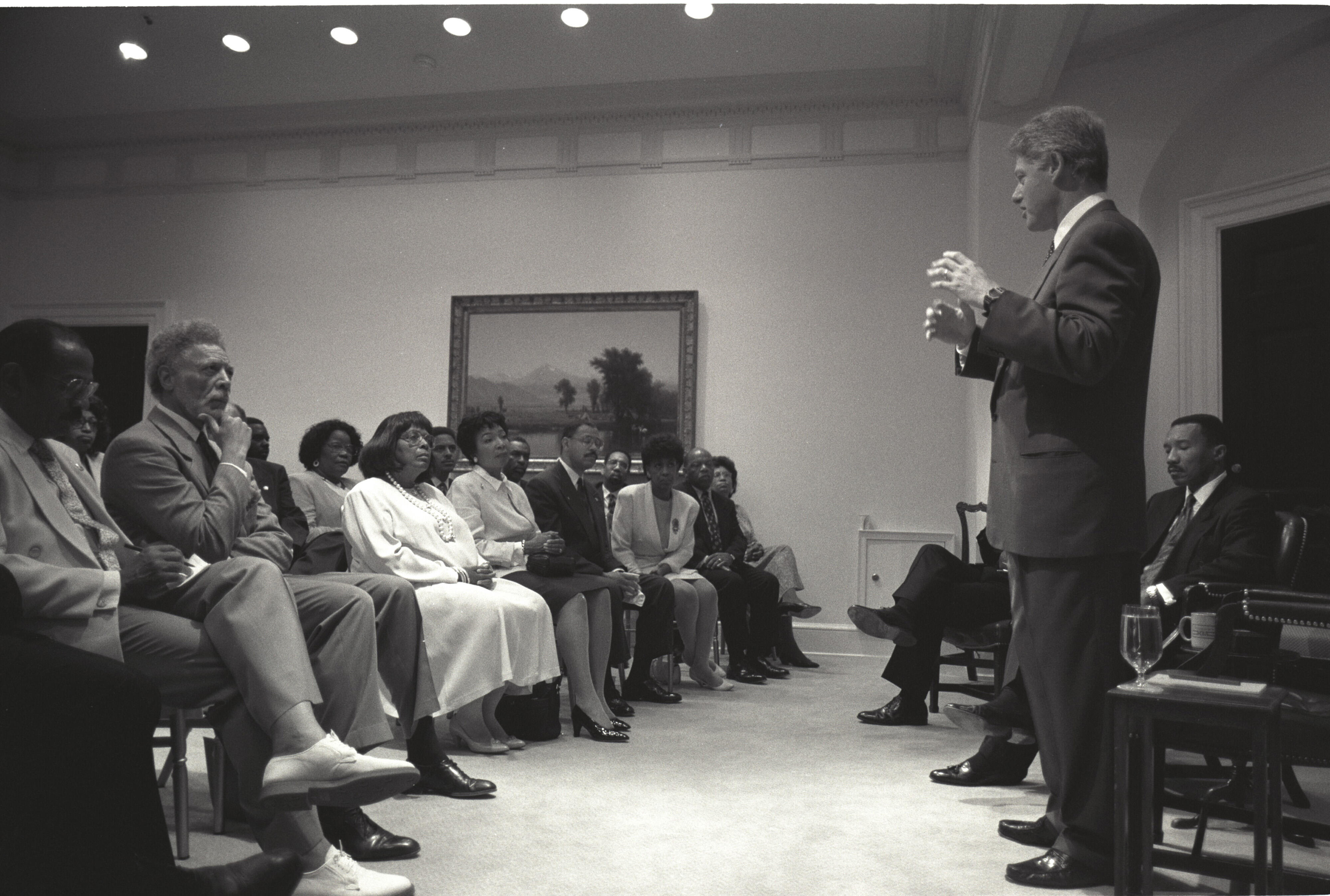 Black and white photograph. President Clinton stands while addressing a sitting audience of member of the Congressional Black Caucus.
