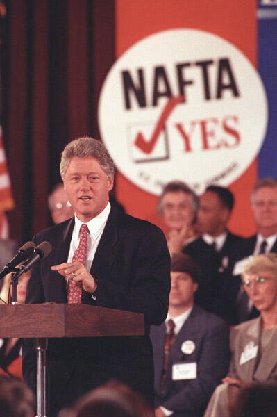 President Clinton stands behind a podium. A large sign in the background reads "NAFTA, Yes." People stand in the background.
