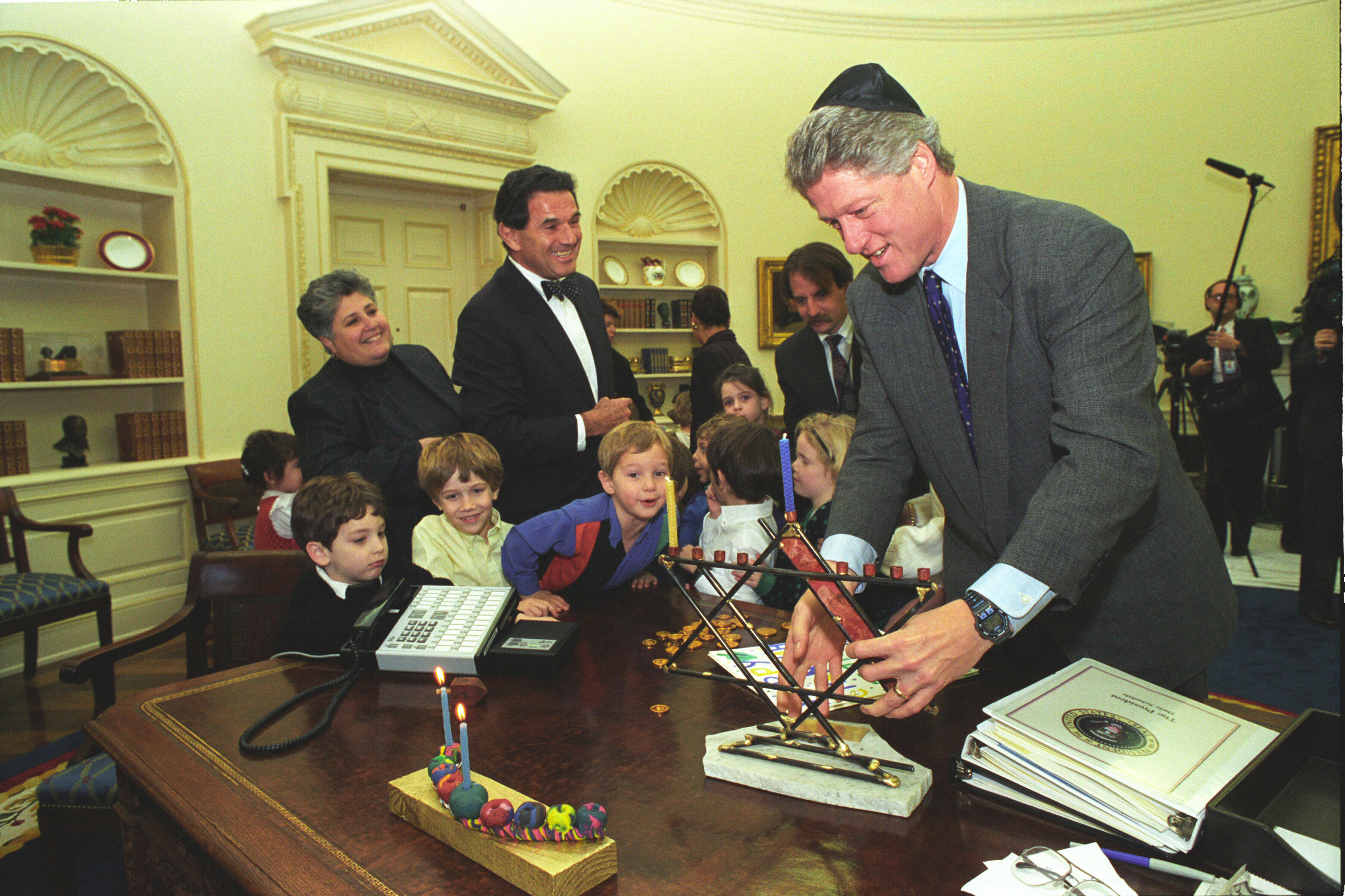 President Clinton places a menorah on the resolute desk in the Oval Office. President Clinton wears a yarmulka. Children lean over desk.