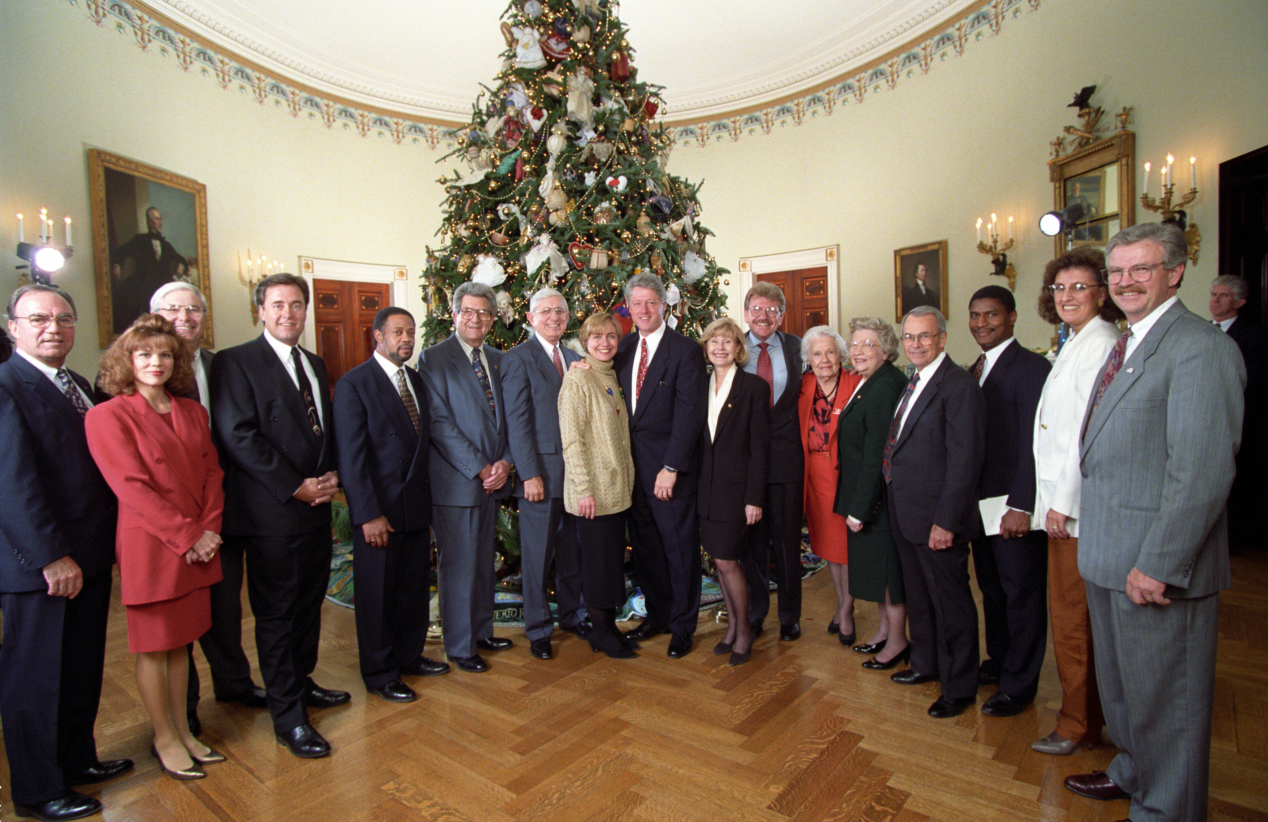 Fifteen people can be seen standing while posing for a photograph with President Clinton and the First Lady in front of a decorated Christmas tree.