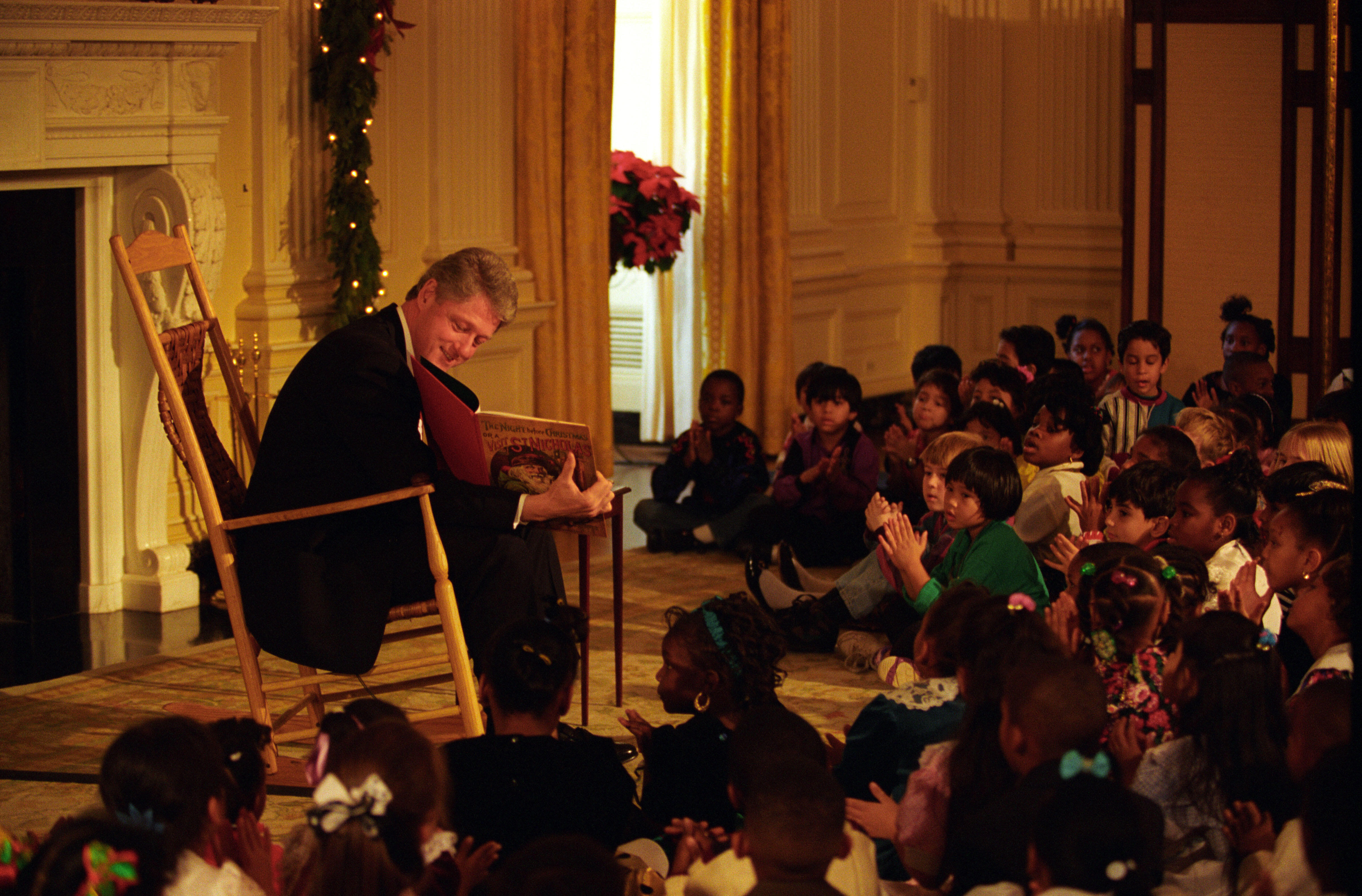 President Clinton sits in a wooden rocking chair and reads to a group of children. President Clinton holds a book. The group of children sit on the floor.