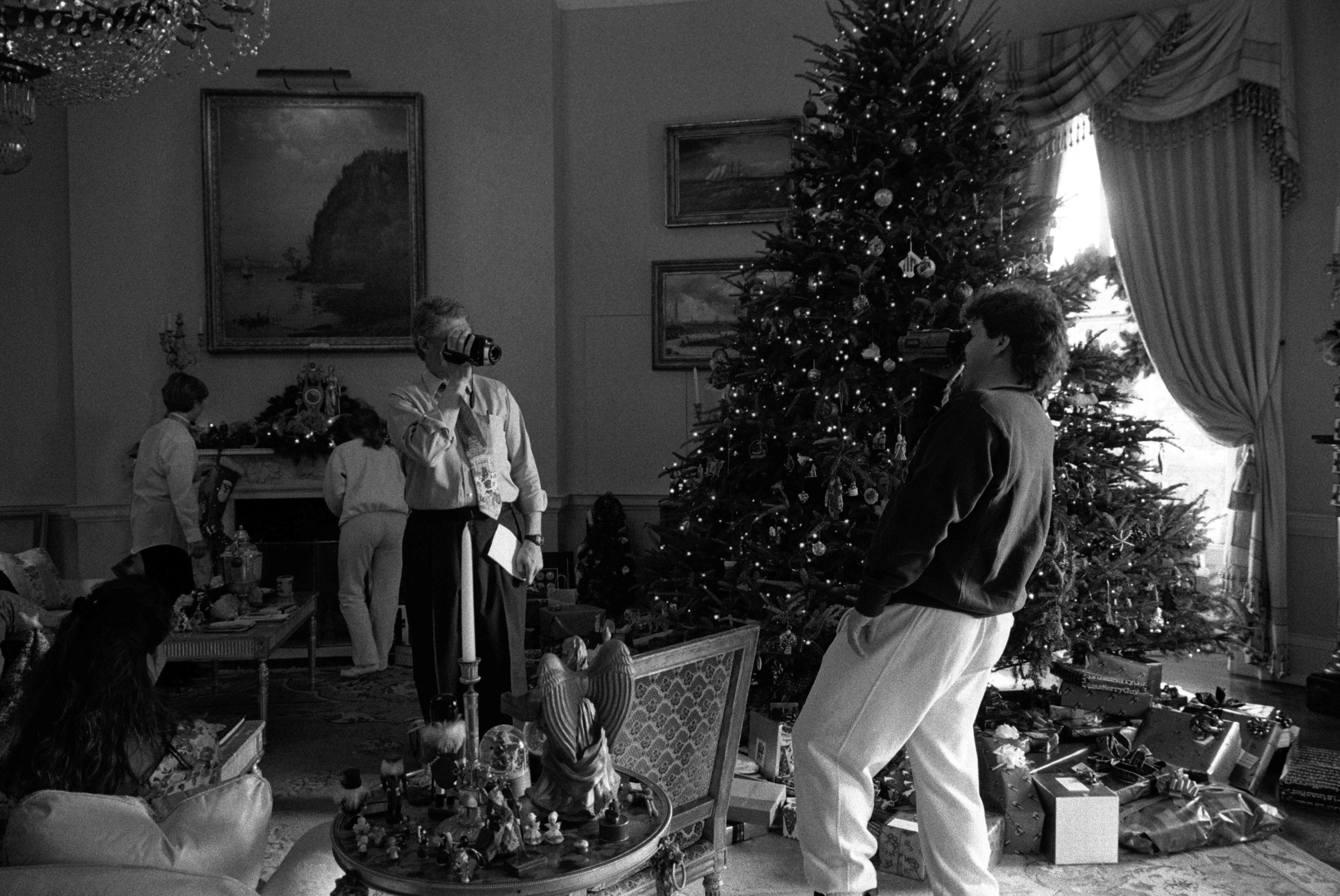 Black and white photograph. President Clinton and and his brother, Roger Clinton film each other with video cameras. Others can be seen standing in the background facing toward a fireplace mantel. A decorated Christmas tree can be seen in the back corner of the room.