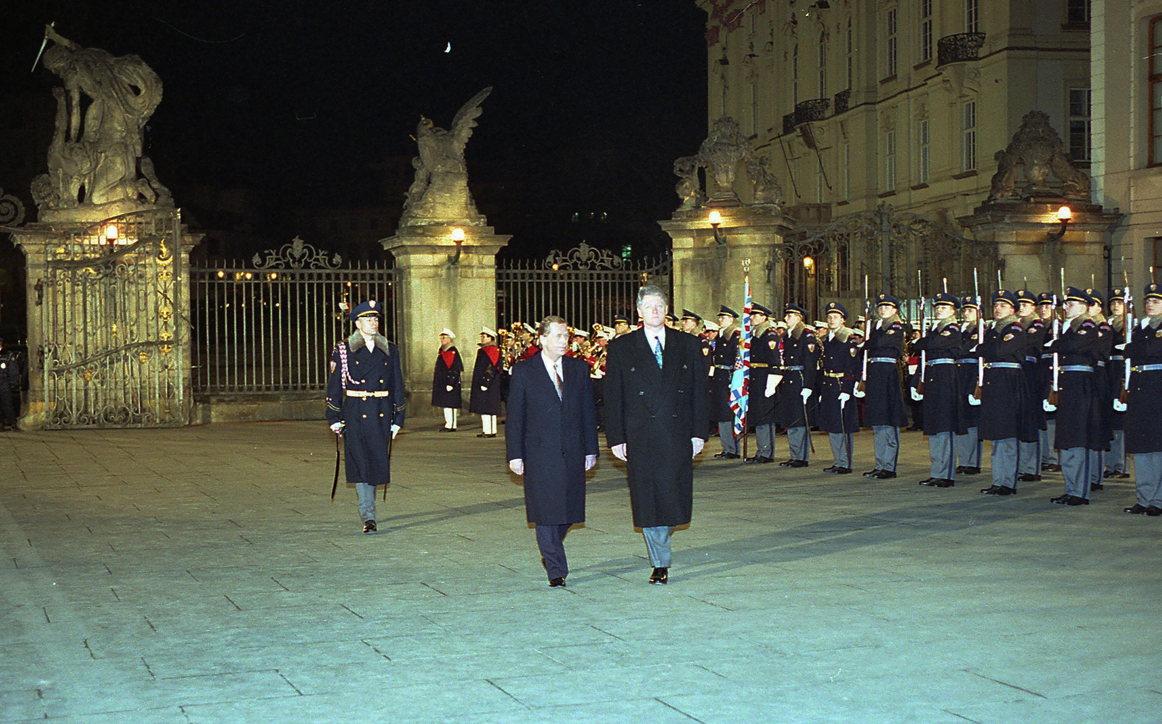 President Clinton and Václav Havel walk through the Matthias Gate in the courtyard of Prague Castle. The courtyard is flanked with members of the Prague Castle Guard standing at attention. The Photograph is taken and night. One of the "Giants" statues of the gate may be seen in the background.