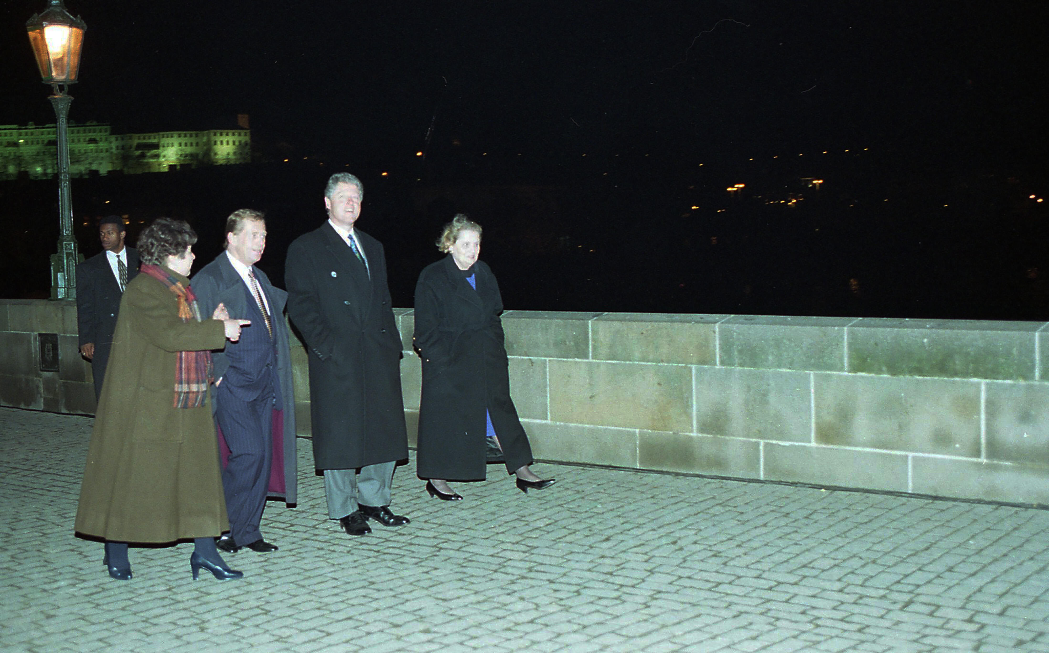 Photograph taken at night. President Clinton, President Vaclav Havel, Ambassador Madeleine Albright, President Clinton's Security Staff, and an unknown woman all wall across a stone bridge. They all wear overcoats. The lights of the city of Prague can be seen in the background.