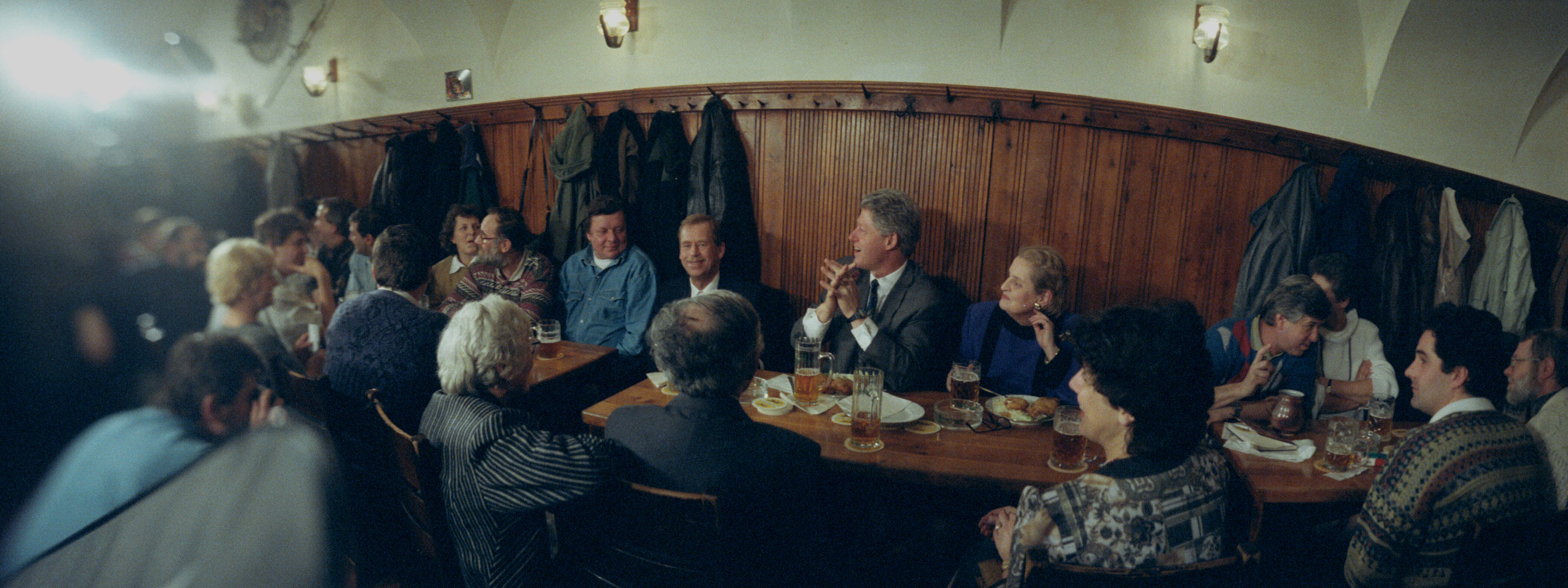 President Clinton, President Václav Havel, Ambassador Madeleine Albright, and at least 15 other unidentified peopl sit at a large table in a pub. The table is full of plates of food and glasses of beer. Coats and jackets hang on the wall behind the table. a photographer can be seen facing President Clinton, taking a photograph. 