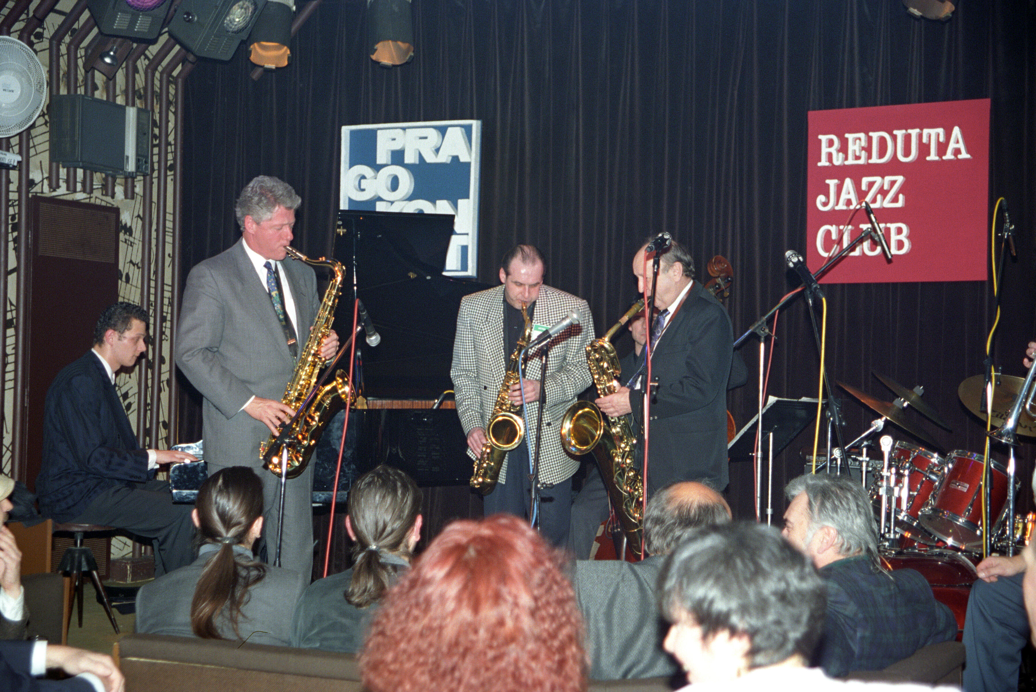 President Clinton stands and plays a brass saxaphone with two other people also standing and playing saxaphones on a stage. A man sits playing the piano behind President Clinton. Miphone stands and a large black curtain can also be seen in the background.