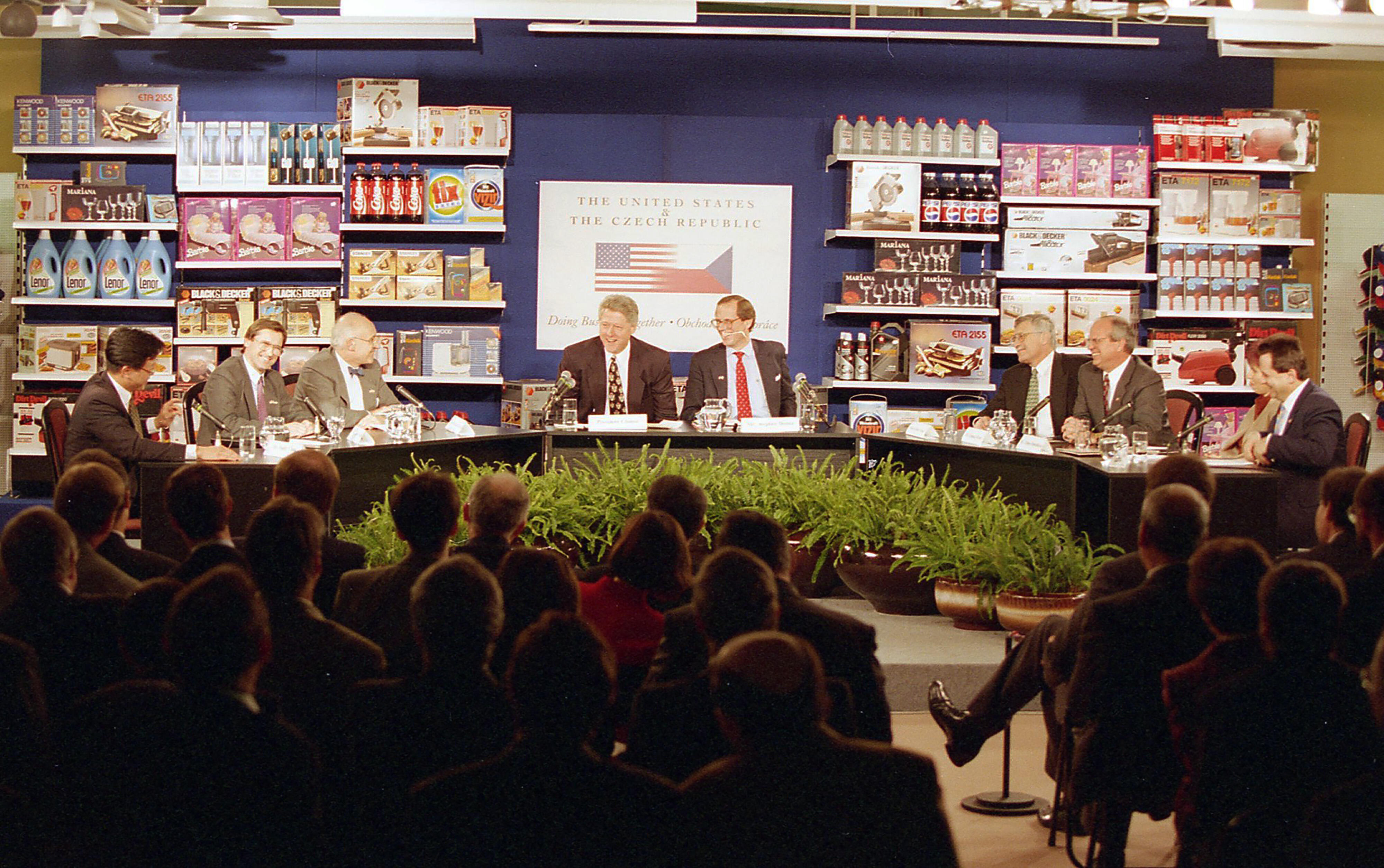 President Clinton sits at a table among seven panel participants. The panel sits infront of a wall of products (like Coca-Cola, Pepsi, wine glasses, toaster, laundry detergent, circular saw, etc.) sold at the K-Mart. A crowd of attendess can be seen in the foreground of the photograph, facing the panel participants. The Panel table is decorated with green plants.
