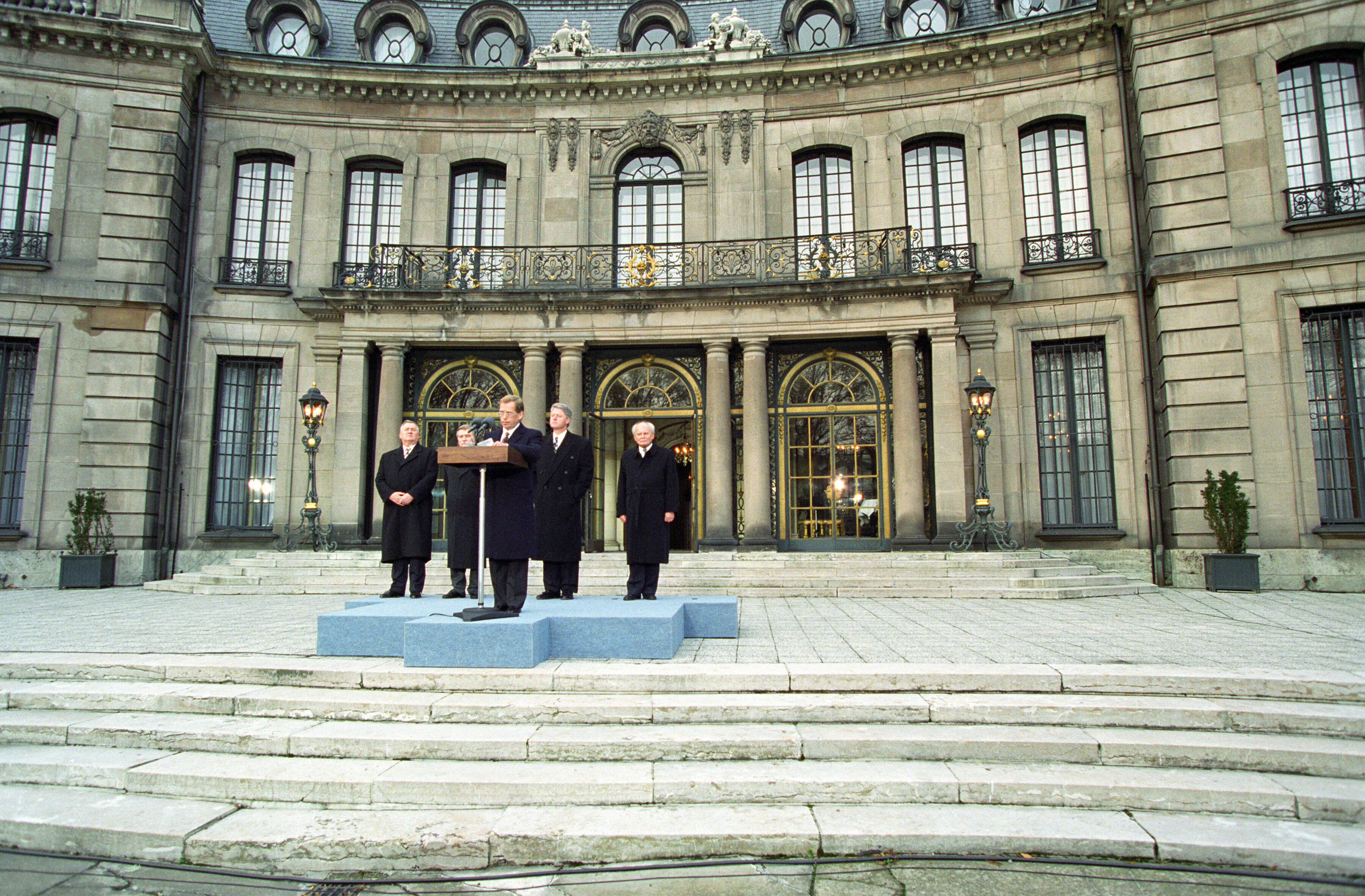President Václav Havel stands behind a podium on a light blue stage. The stage sits on stone stairs. President Clinton, President Michal Kovac of Slovakia, President Lech Walesa of Poland, and President Arpad Goncz of Hungary stand behind President Havel. Petschek Villa, an opulent neo-baroque building with gilded doors and columns can be seen in the background.