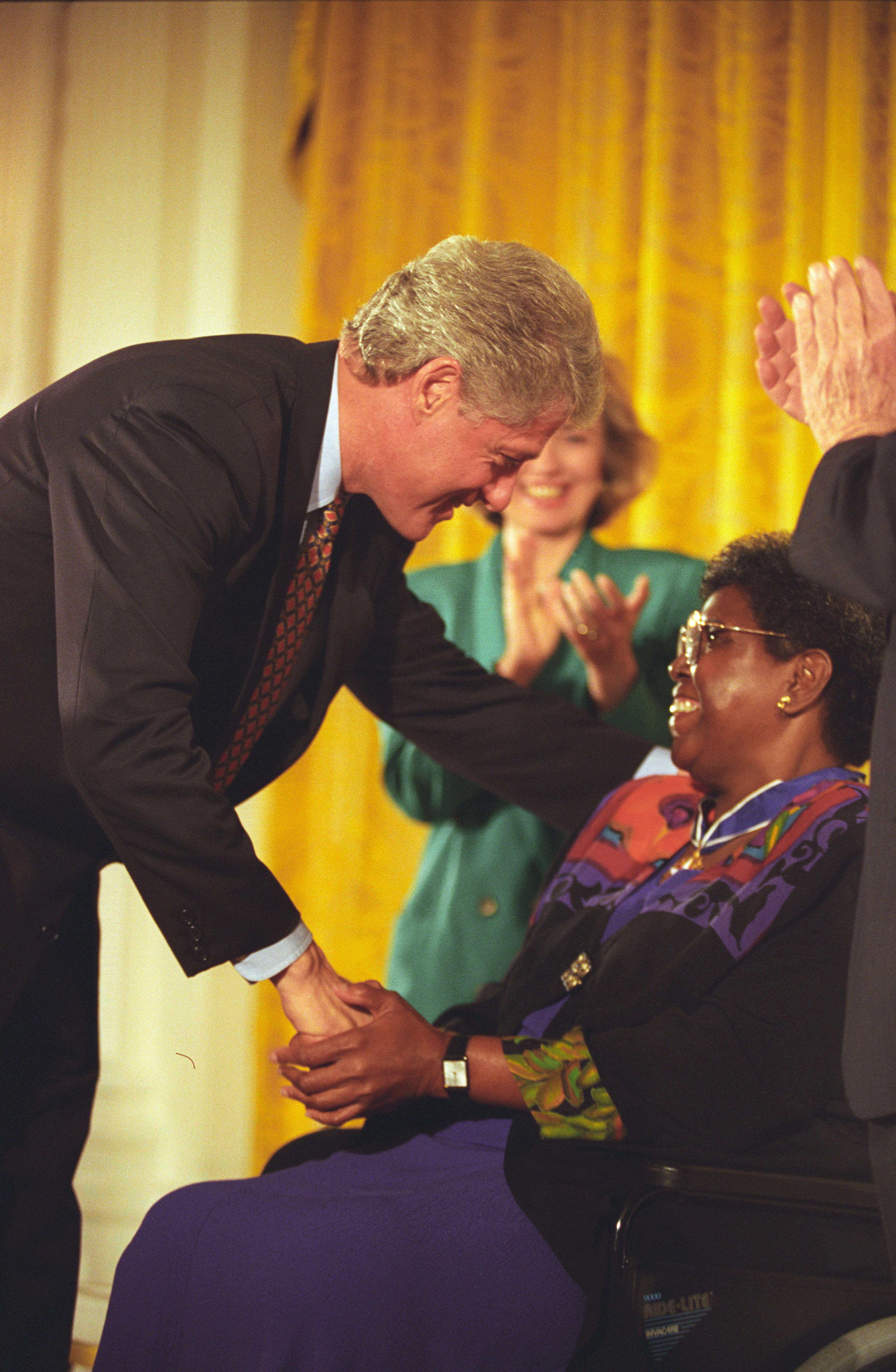 President Clinton leans over to shake hands with Barbara Jordan who is sitting down.