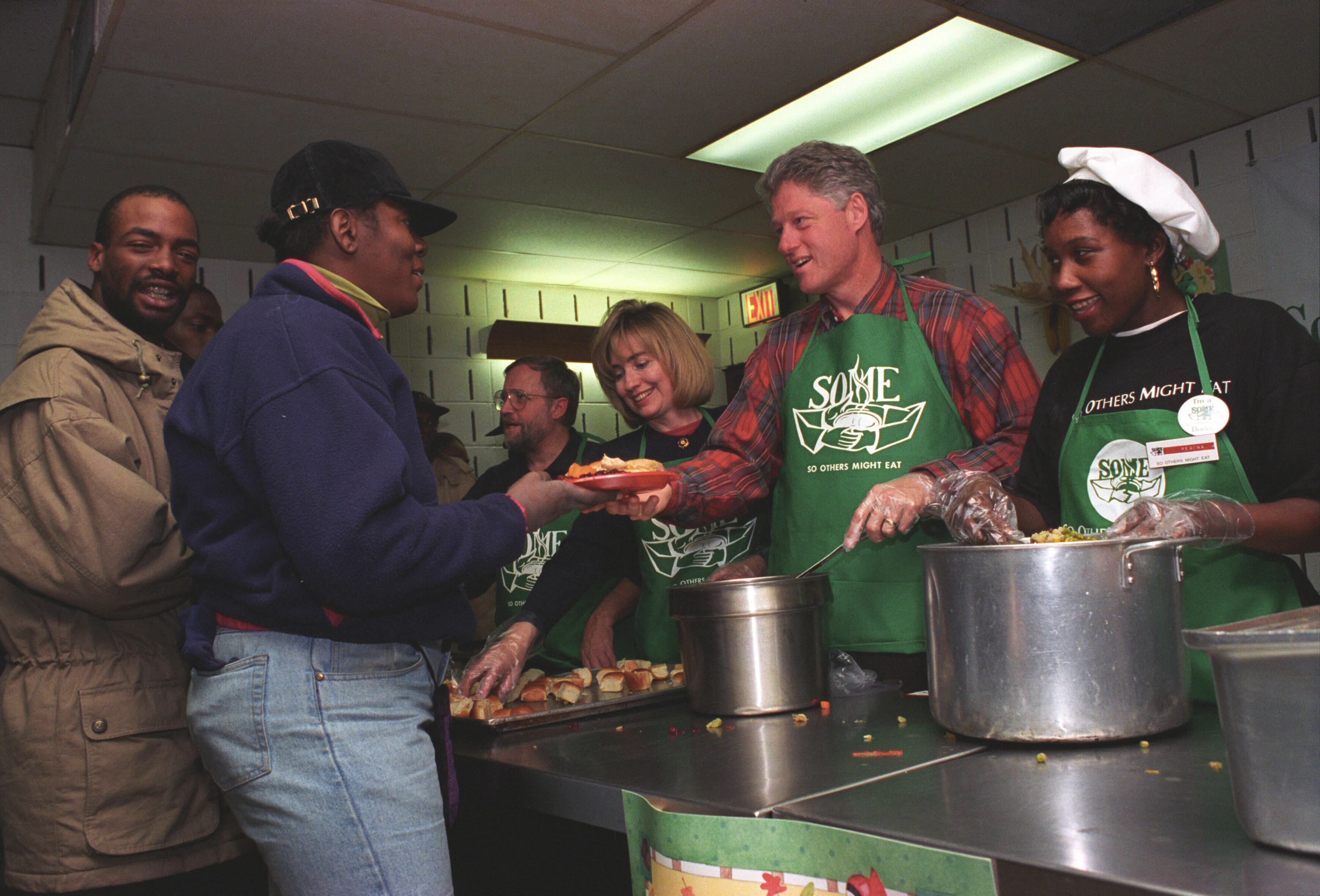 President Clinton and First Lady Hillary Rodham Clinton serve a Thanksgiving meal. The President and First Lady wear aprons and stand next to two other kitchen volunteers. They serve two men standing in line. 