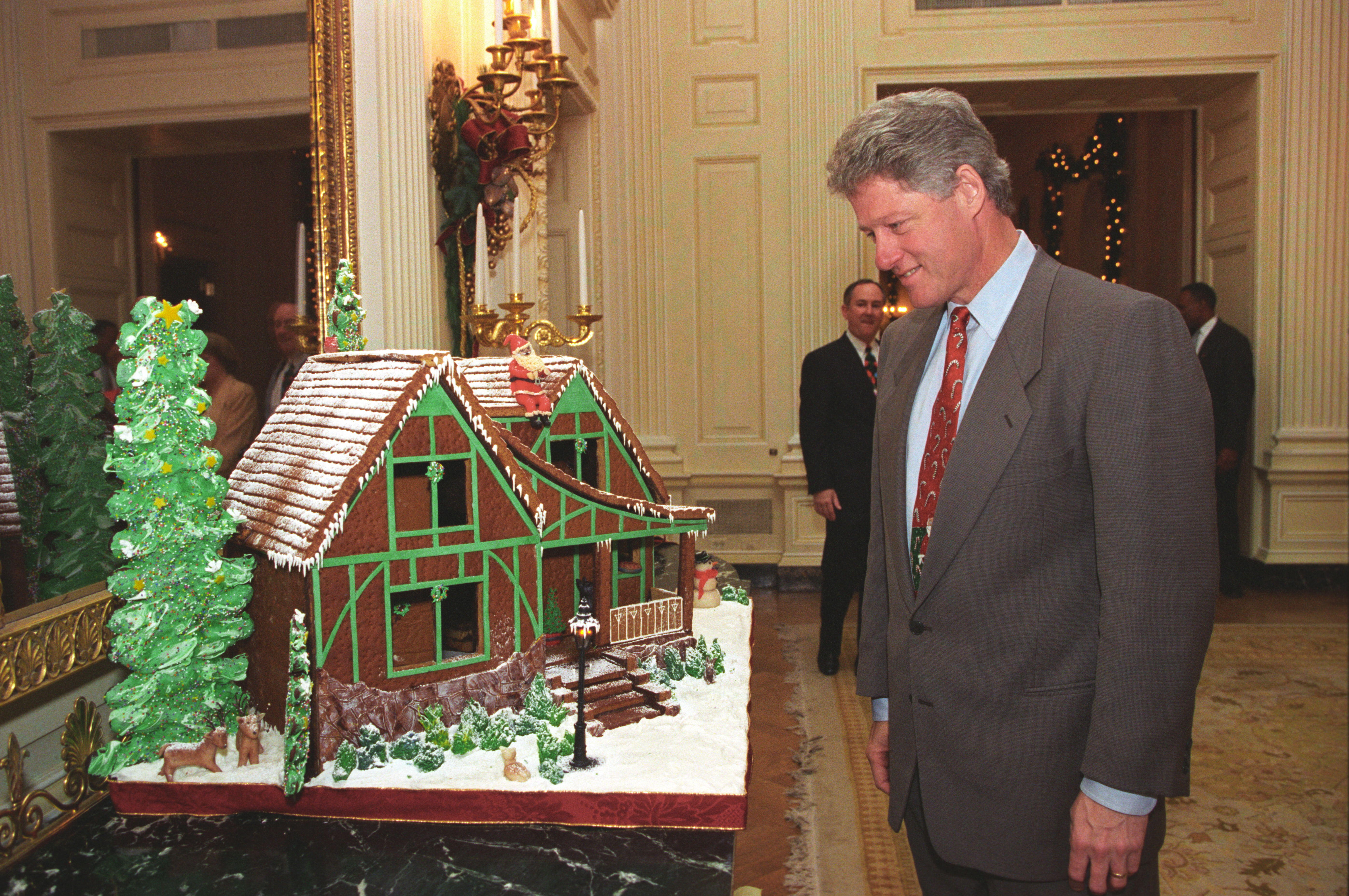 President Clinton stands and views a gingerbread house displayed on a table. Two men can be seen standing in the background.