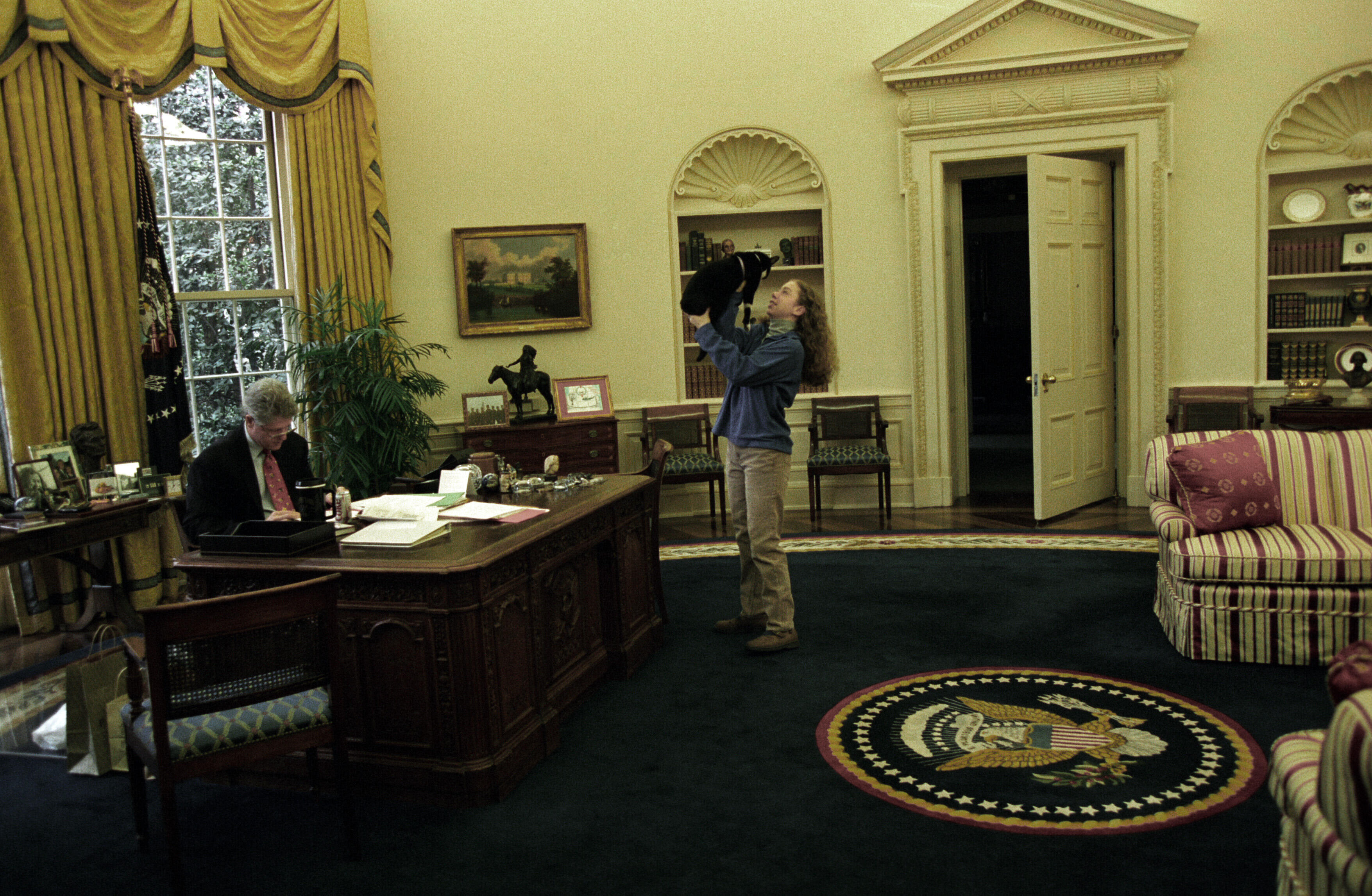 Chelsea Clinton holds Socks the Cat above her head while President Clinton works behind the Resolute Desk in the Oval Office of the White House