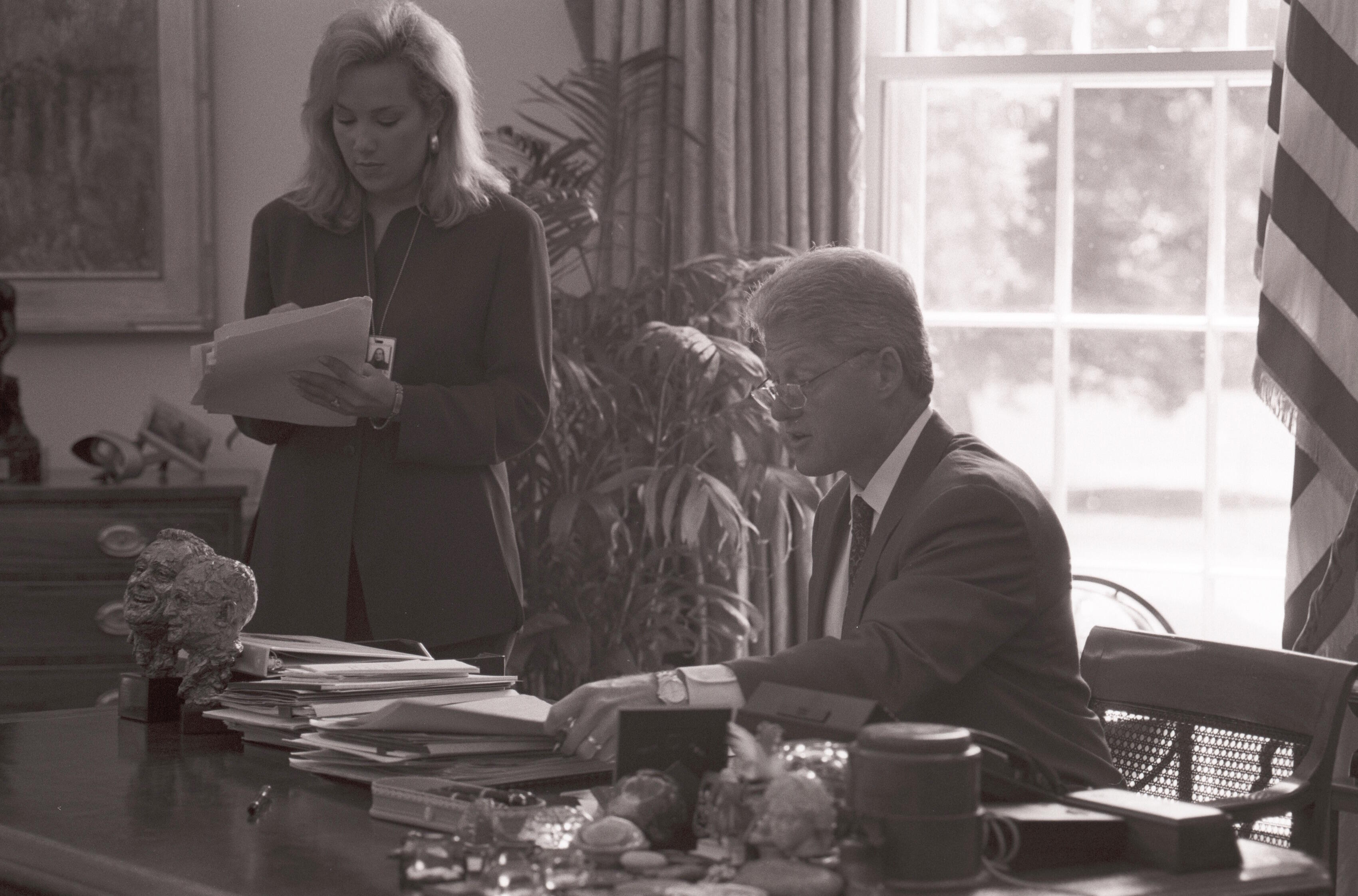 Black and white photograph. President Clinton meets in the White House Oval Office with the Director of the Office of Scheduling, Stephanie Streett. President Clinton sits behind the resolute desk. Stephanie Streett stands holding a stack of papers.
