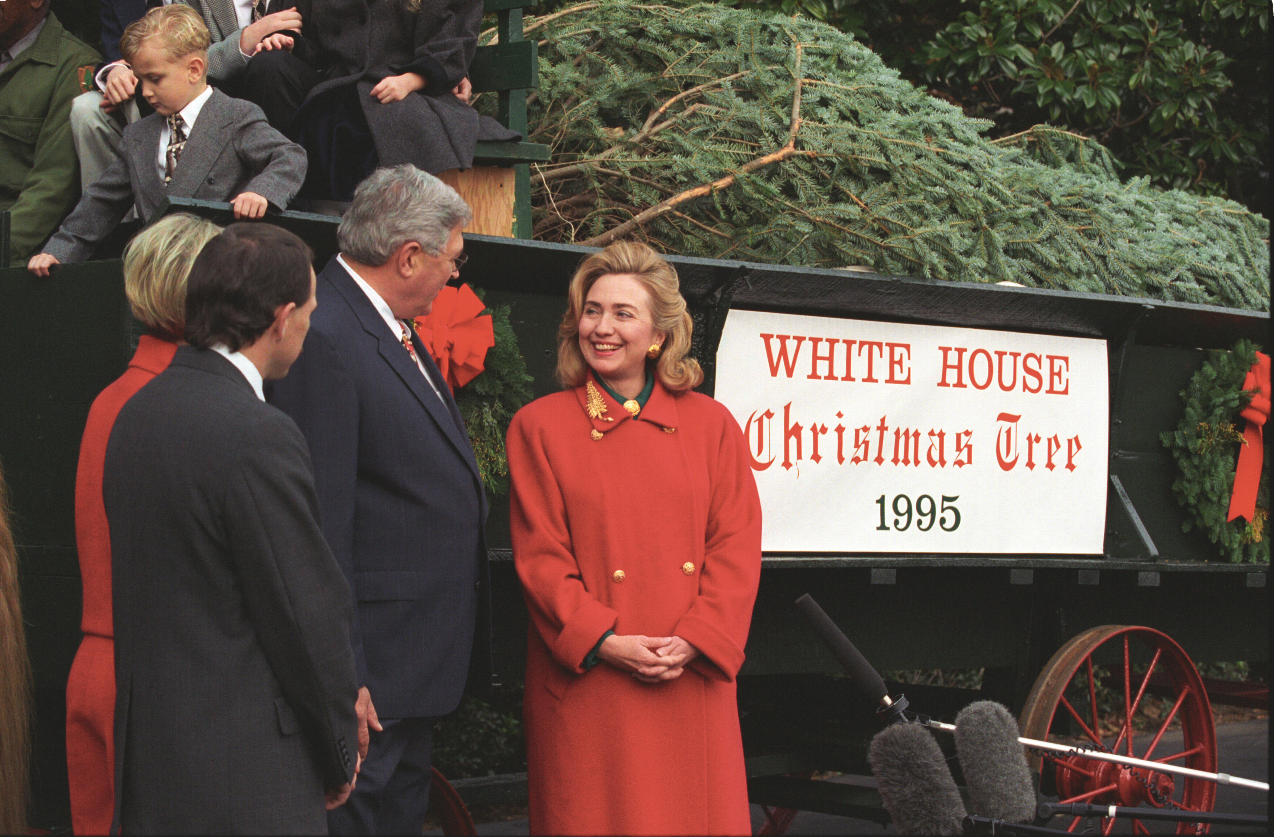 First Lady Hillary Rodham Clinton stands facing three other people in front of a green horse drawn wagon. The wagon carries the White House Christmas Tree. A boy sits on the wagon. 