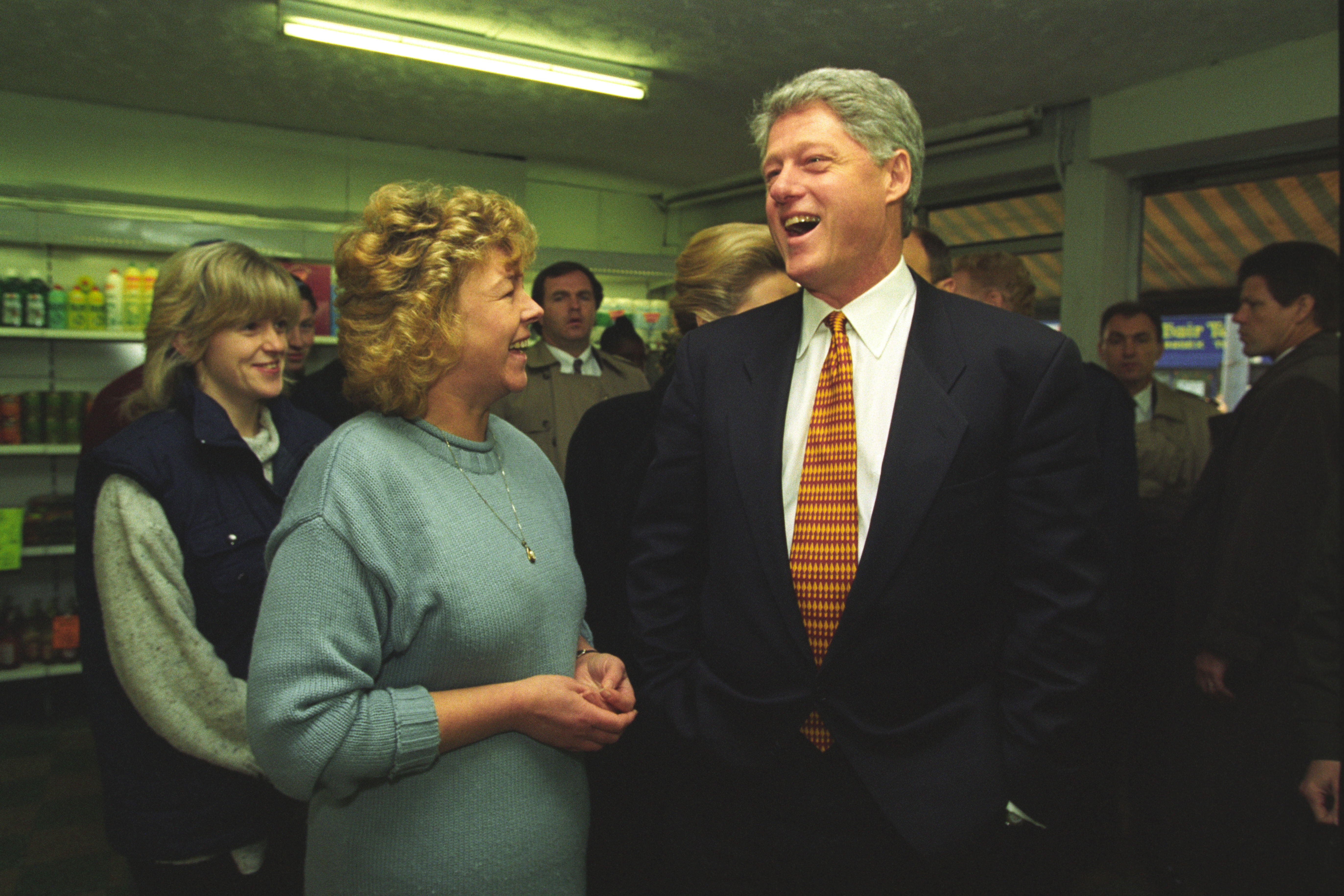 Victoria "Violet" Clarke stands in a blue sweater, speaking to President Clinton. President Clinton stands with his hands in his pockets. Others people can be seen standing behind President Clinton, including First Lady Hillary Rodham Clinton. 