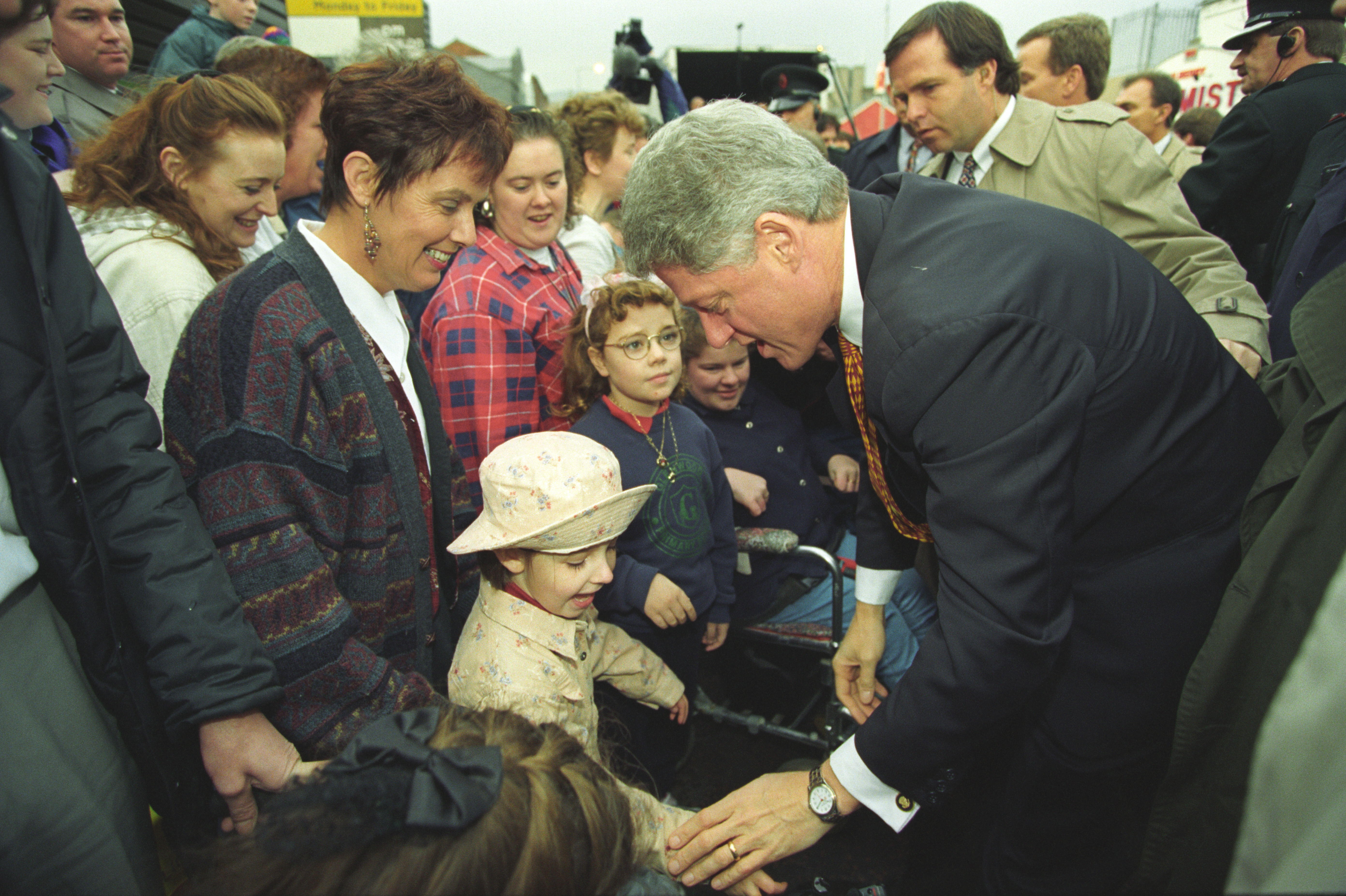 President Clinton leans over to speak with a little girl in a crowd. The girl wears a matching hat and coat. A woman stands behind the girl. 