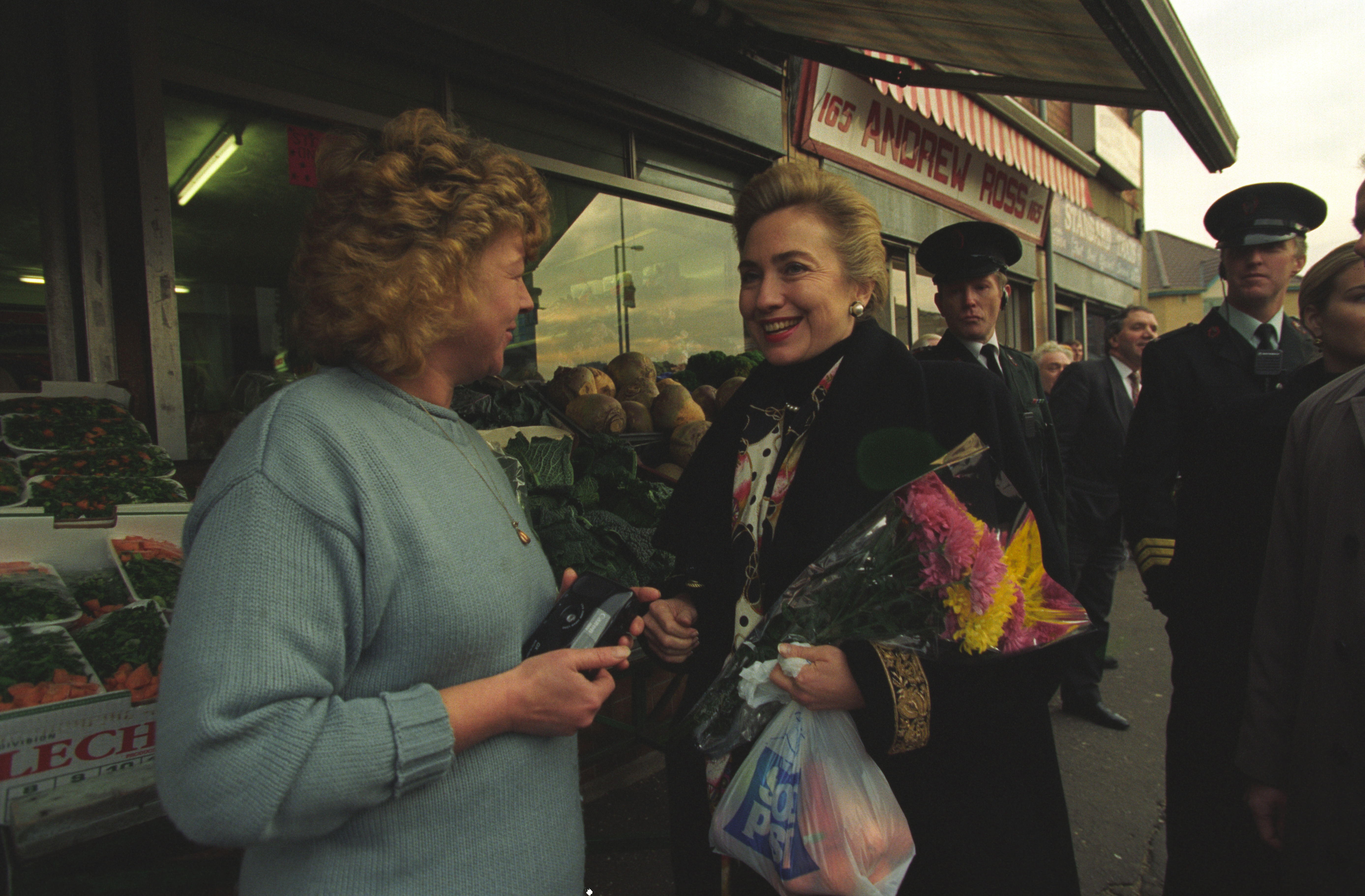 First Lady Hillary Rodham Clinton stands holding a bouquet of flowers, speaking with Victoria "Violet" Clarke. Clarke holds a camera in her hands and wears a light blue sweater. Two uniformed policemen stand in the background.