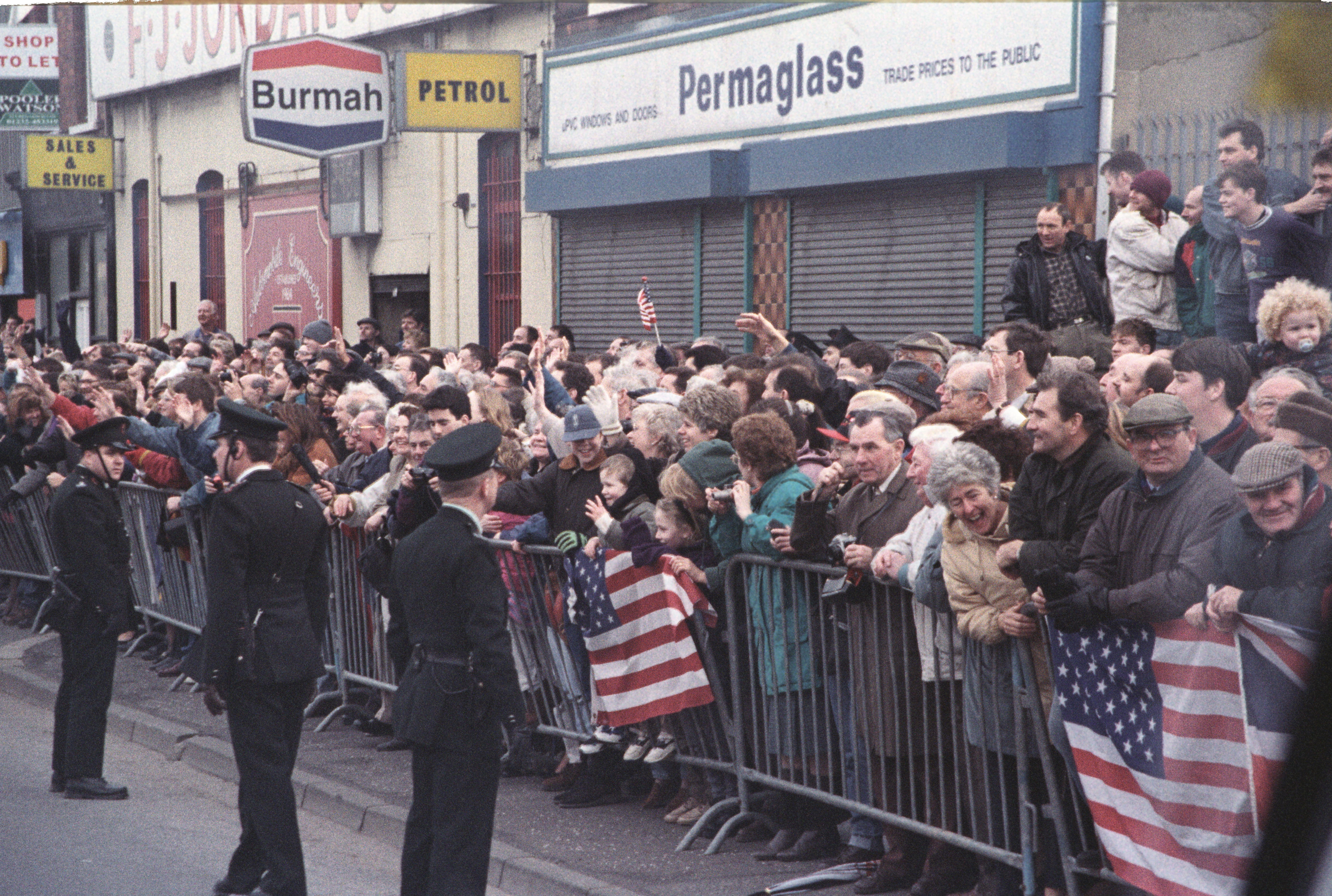 A crowd of people stand behind metal baricades and wave United States flags. Three police in black uniforms stand on the other side of the crowd behind the baricade. 