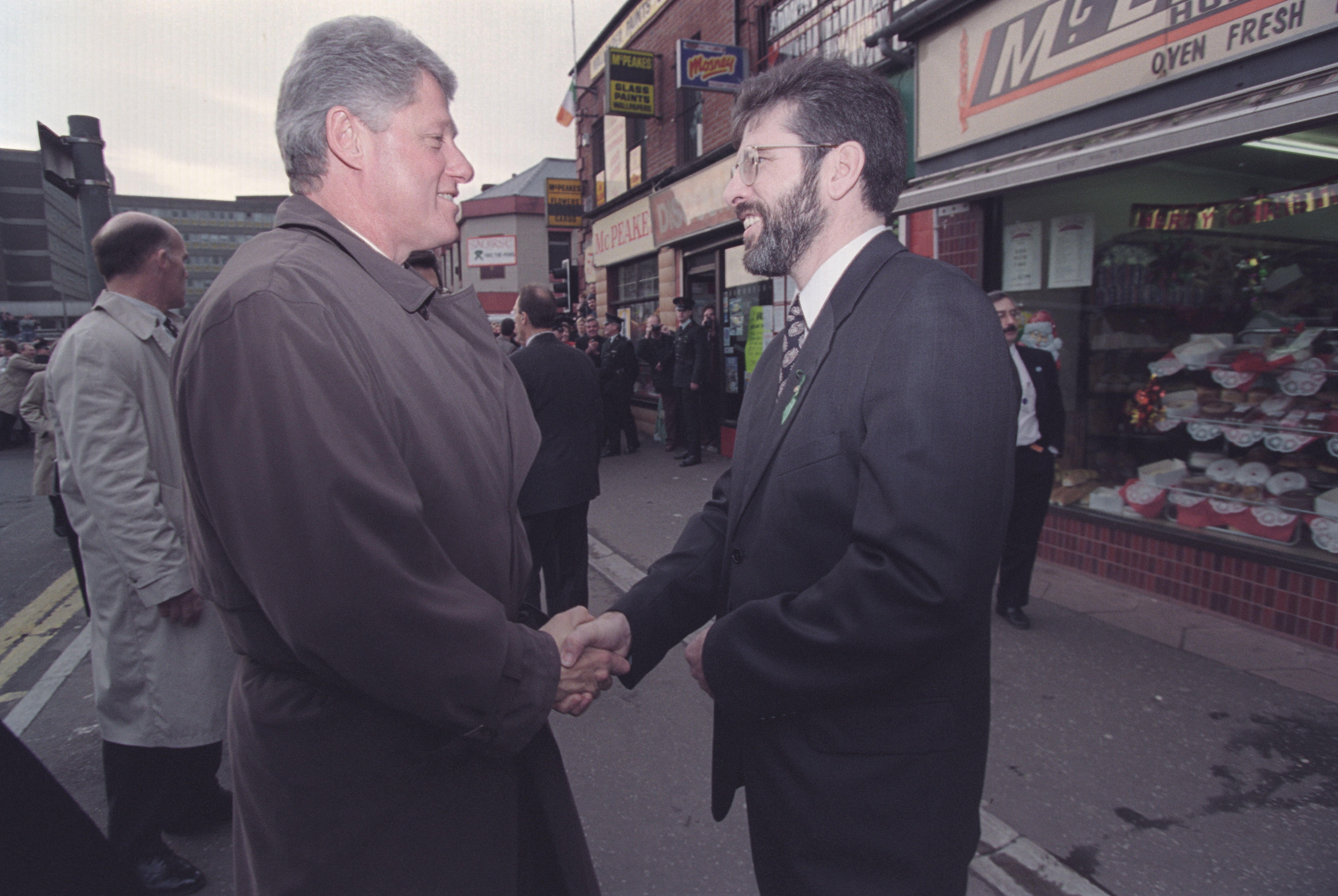President Clinton shakes hands with Sinn Fein in the street. Both men are smiling.