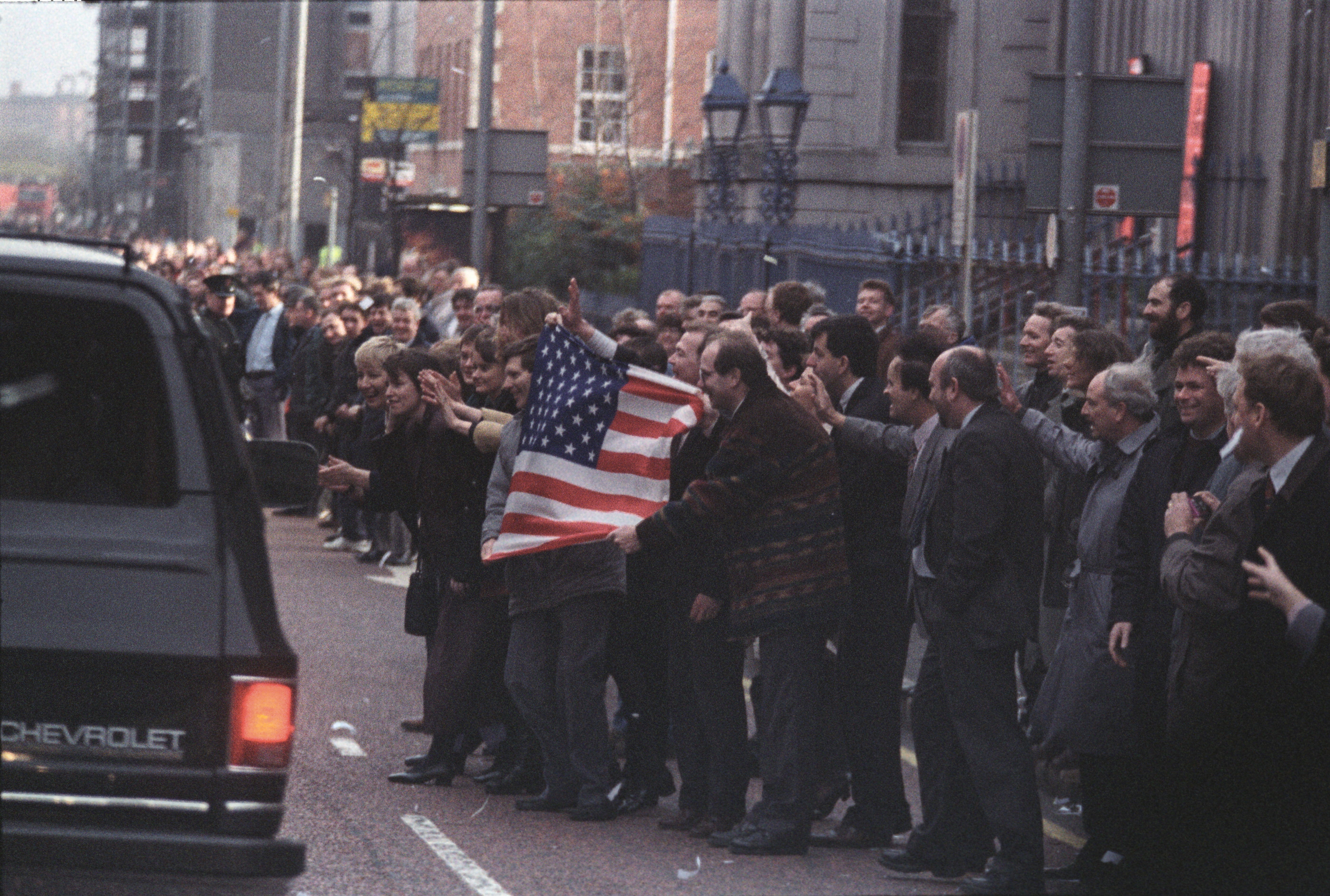 A crowd stands in the street, waving an United States flag at a black Chevorrolet Suburban. People in the crowd wave and clap.