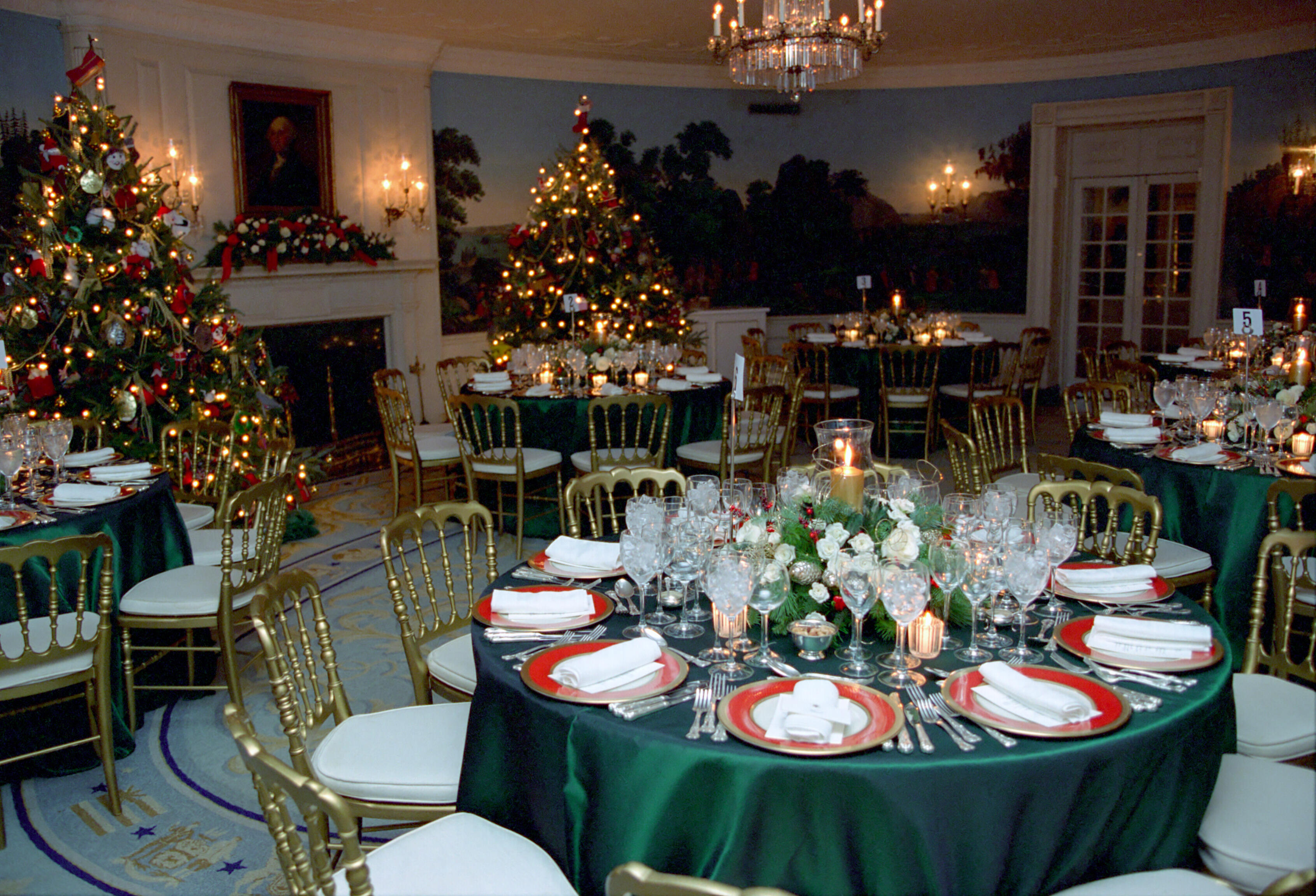 Tables are set for the White House Christmas Dinner Party in the State Dining Room. Tables are covered with green table cloths. The chairs are gold with white seat covers. Two lit Christmas trees are in the background.