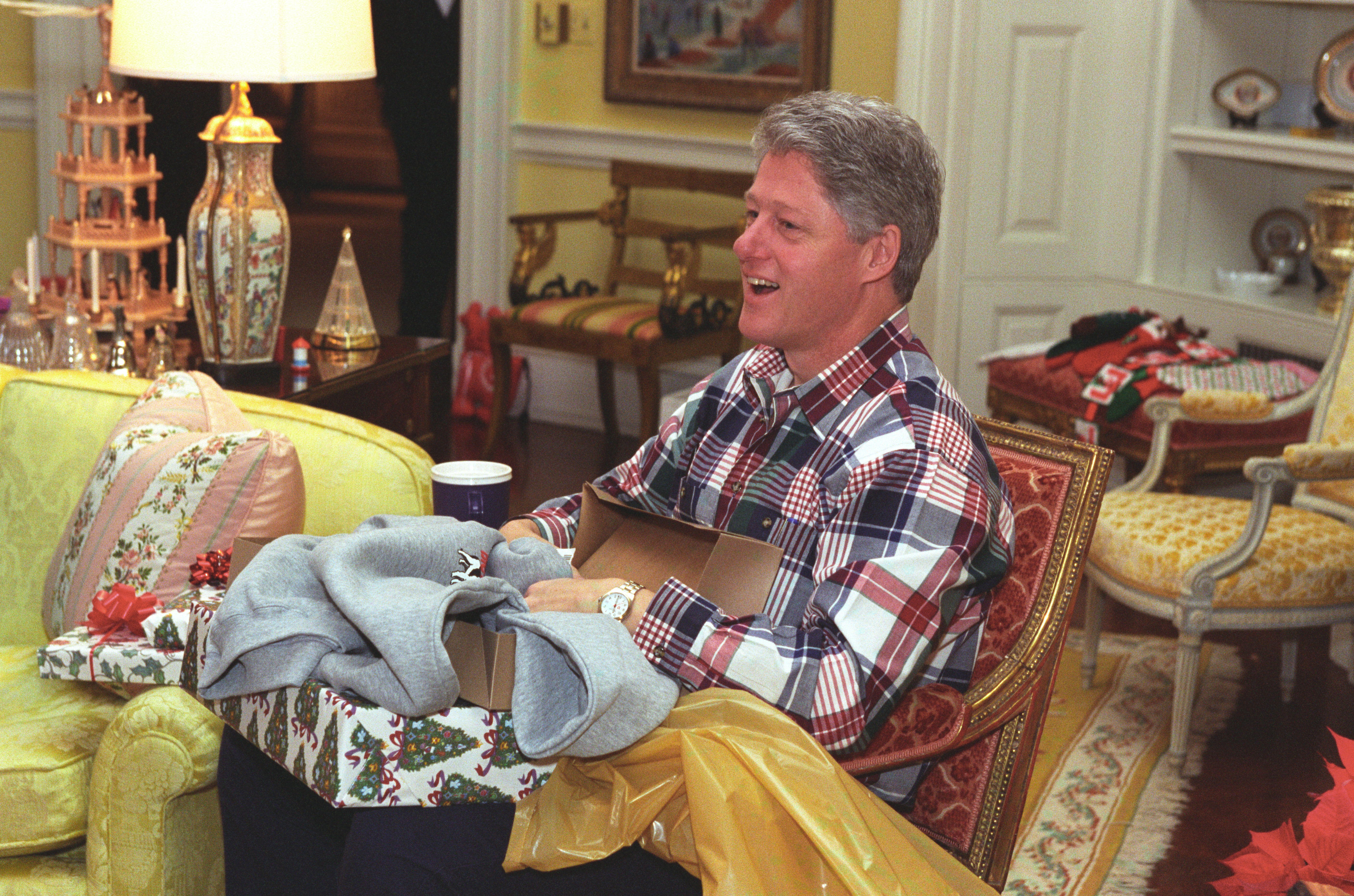 President Clinton sits in a chair with a Christmas present box, wrapping and gift in his lap. 