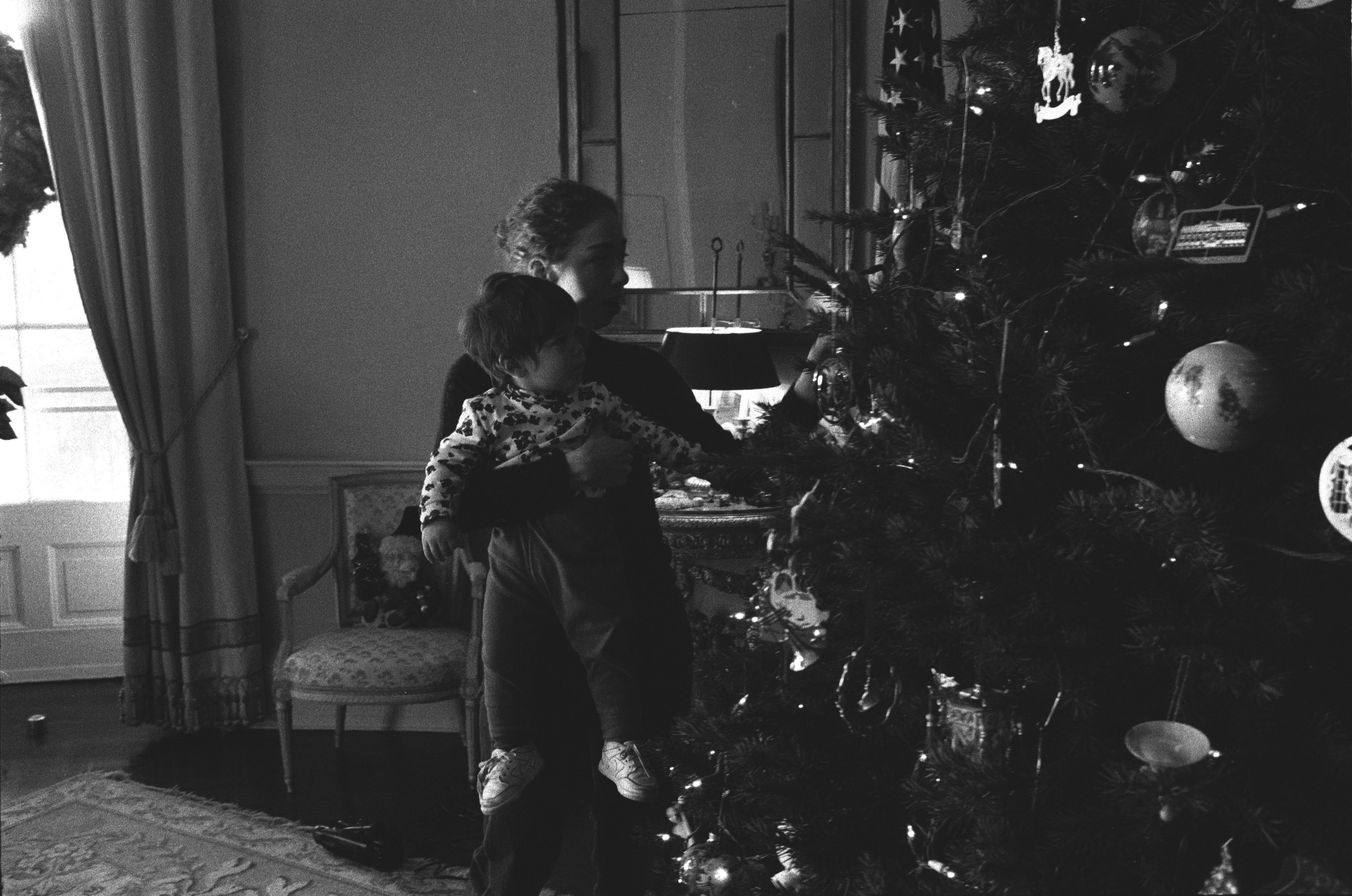 Black and white photograph. Chelsea Clinton holds her cousin, Zach Rodham, as they look at ornaments on the family Christmas tree.