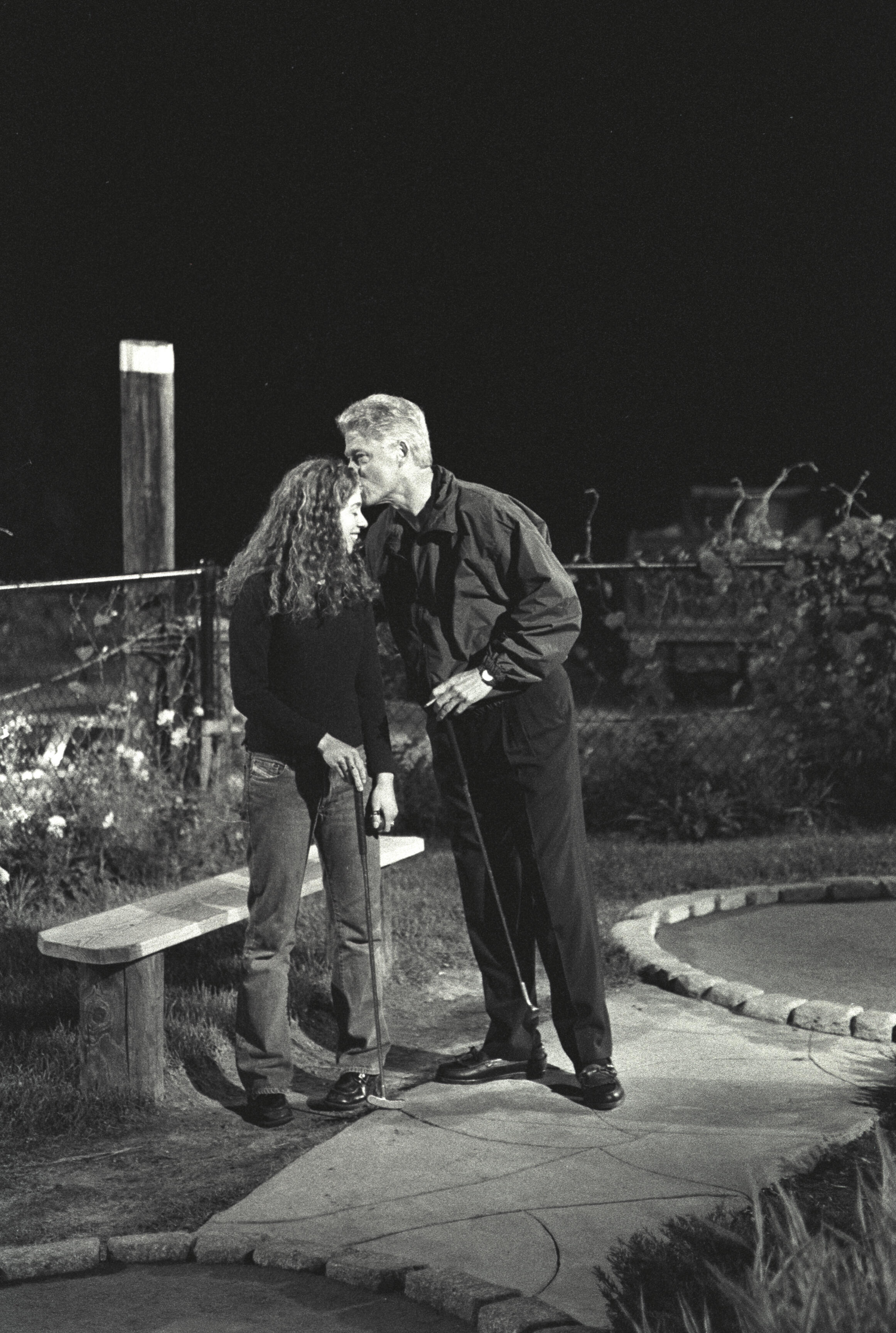 Black and white photograph. President Clinton kisses his daughter, Chelsea Clinton's forehead, while playing mini golf