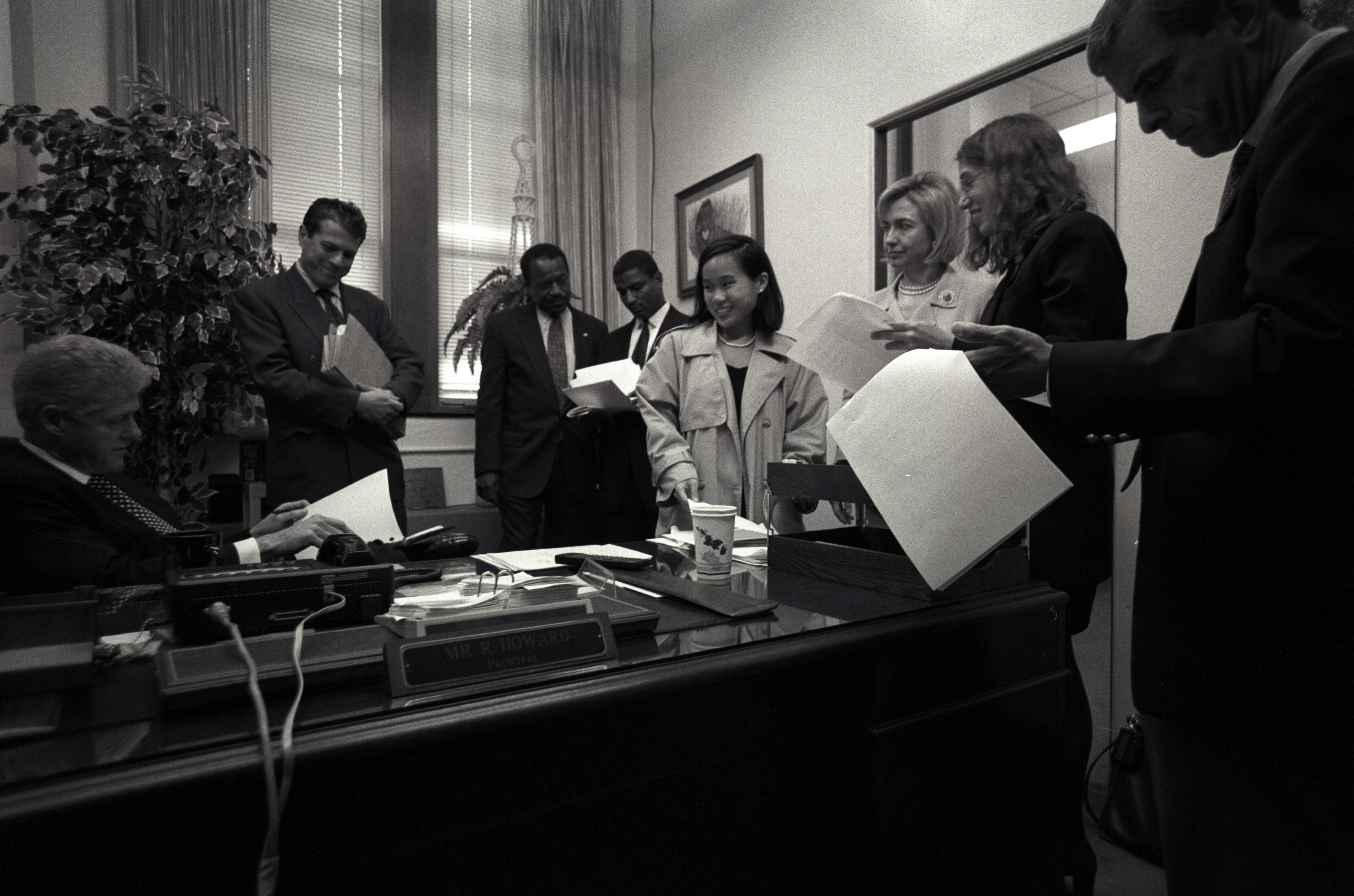 Black and white photo. President Clinton sits at a desk all the others in the room stand